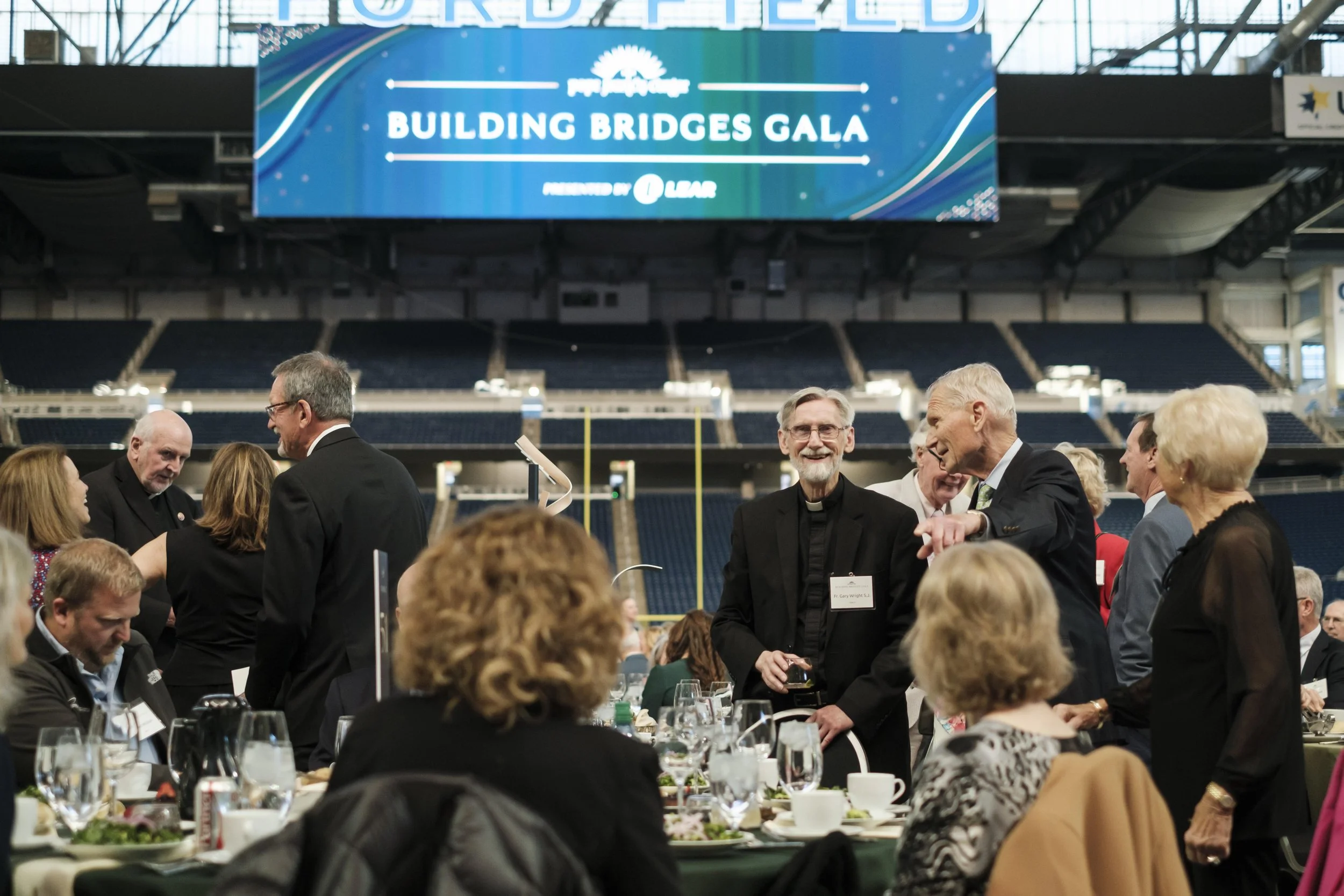 An older man smiles while talking to another man while standing in a crowd during a gala event.