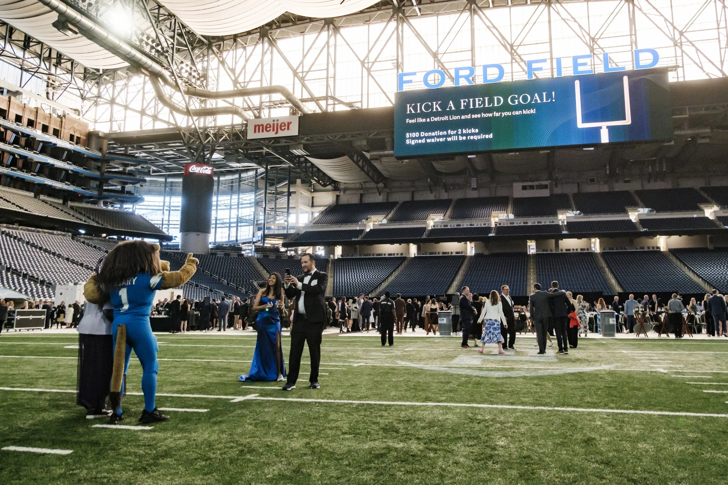 People pose for a photo on Ford Field while wearing black tie attire and standing with the Detroit Lions mascot.