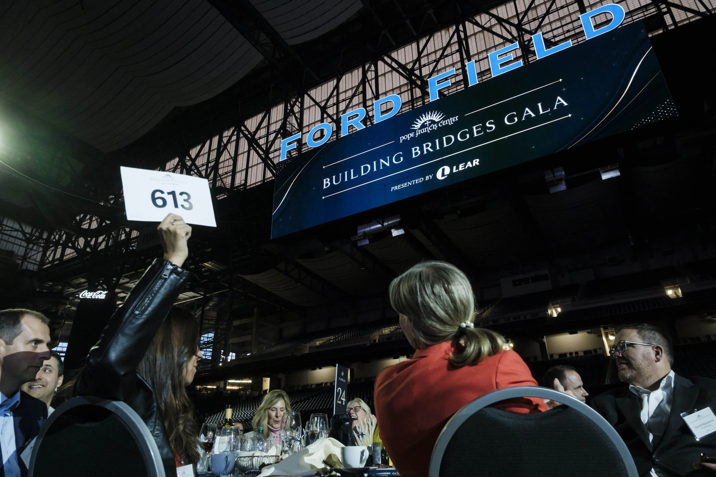 A person holds up a piece of paper with the number 613. They are sitting at a table and illuminated by stage lights.