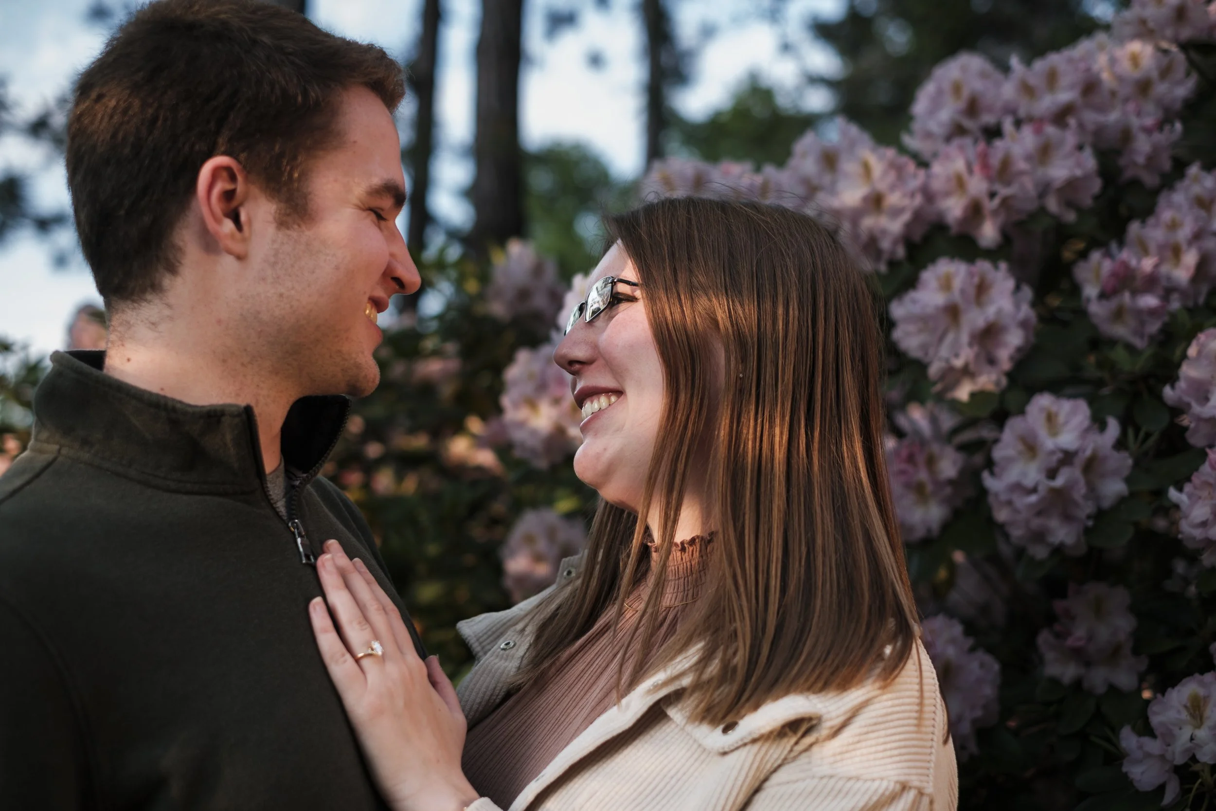 A woman puts her hand on the chest of a man while wearing an engagement ring with a flowering tree in the background.