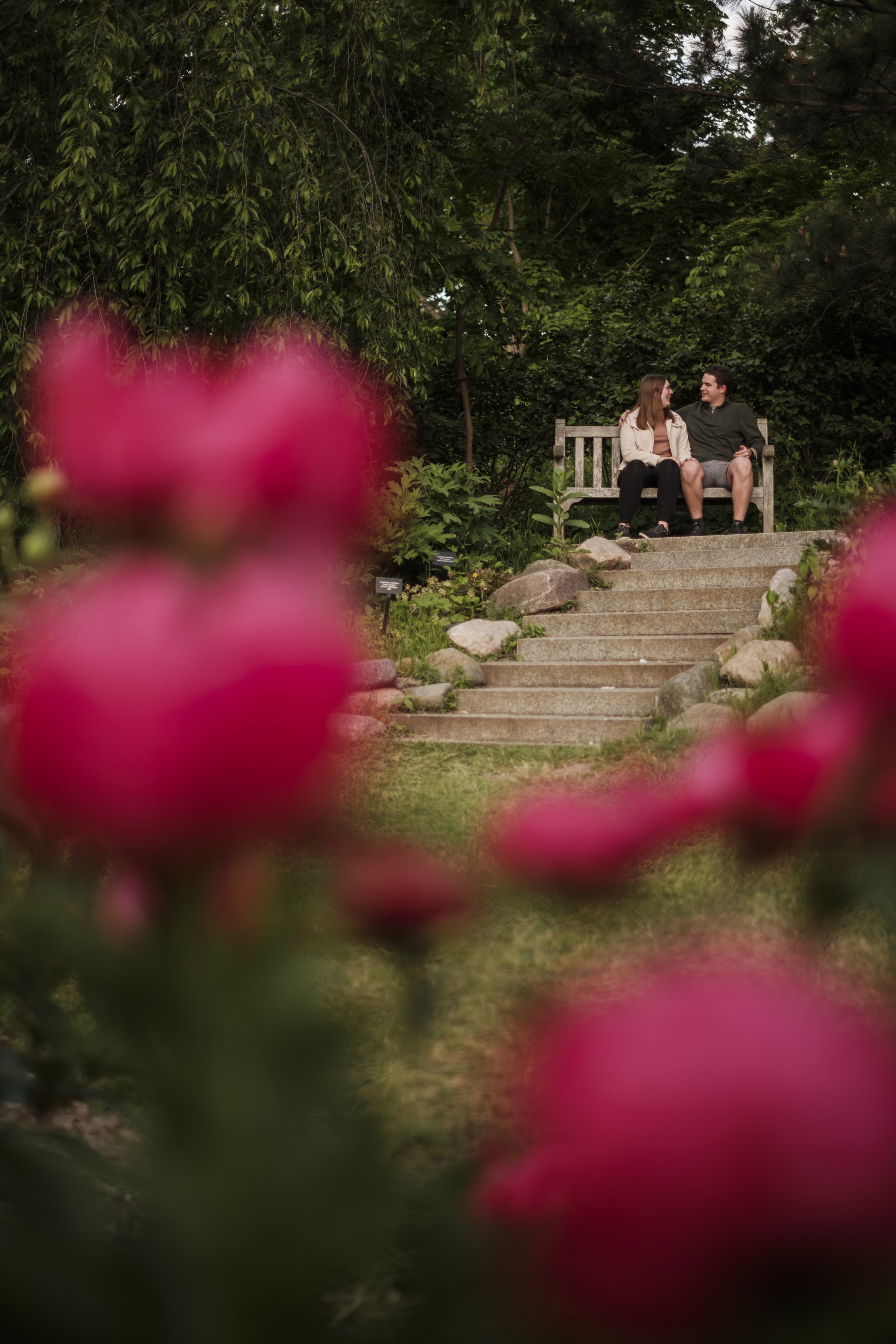 A couple sit on a bench in front of concrete stairs with red blurry flowers in the foreground.