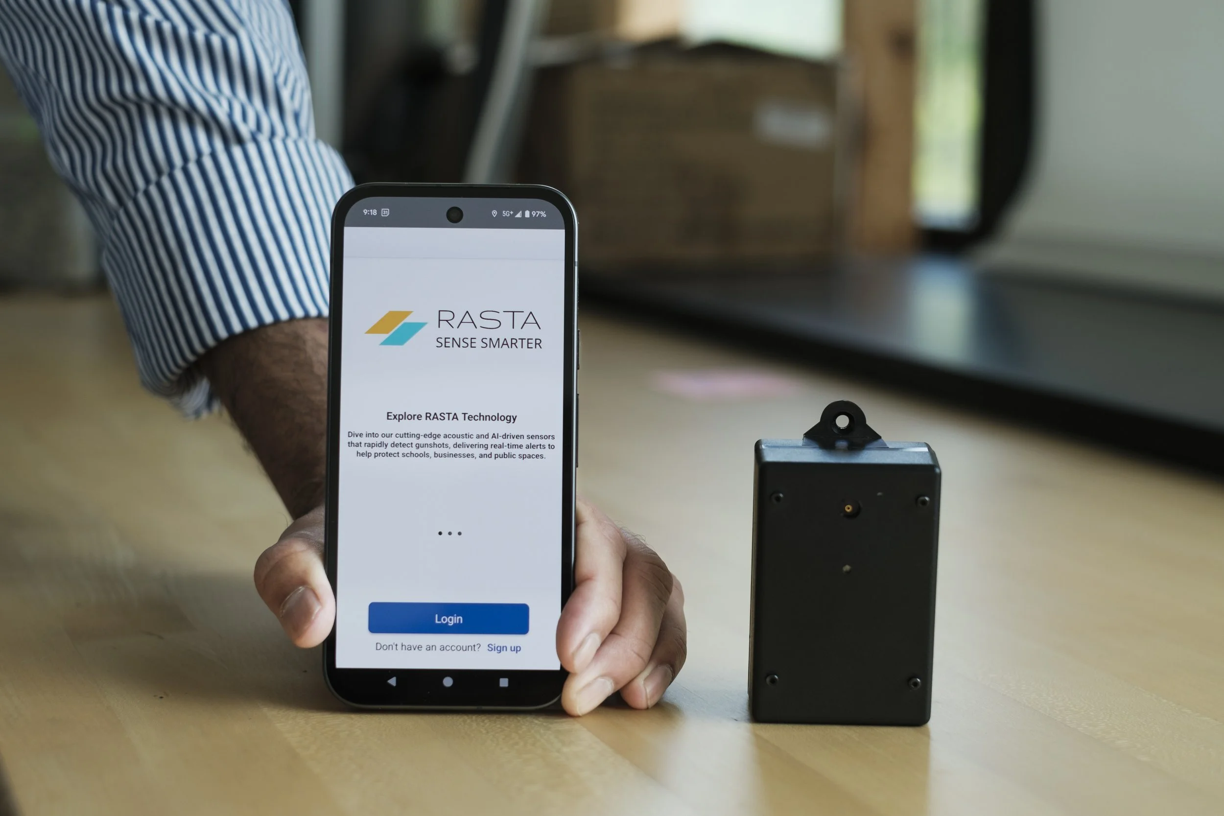 A phone is held up by a hands, while it sits on a table next to a small black device.