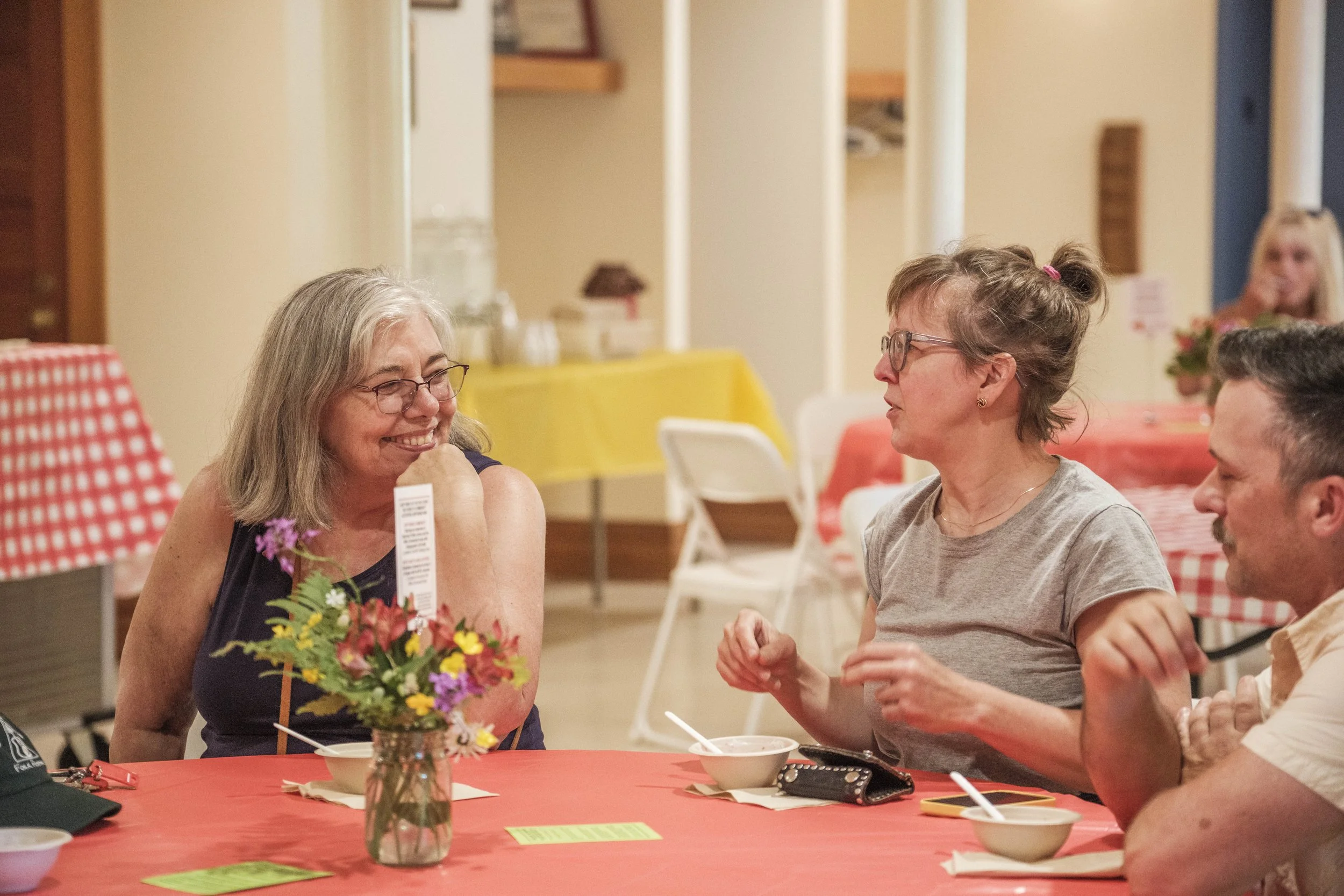 A woman sits at a table and smiles at another woman sitting across from her with a jar of flowers in the foreground..