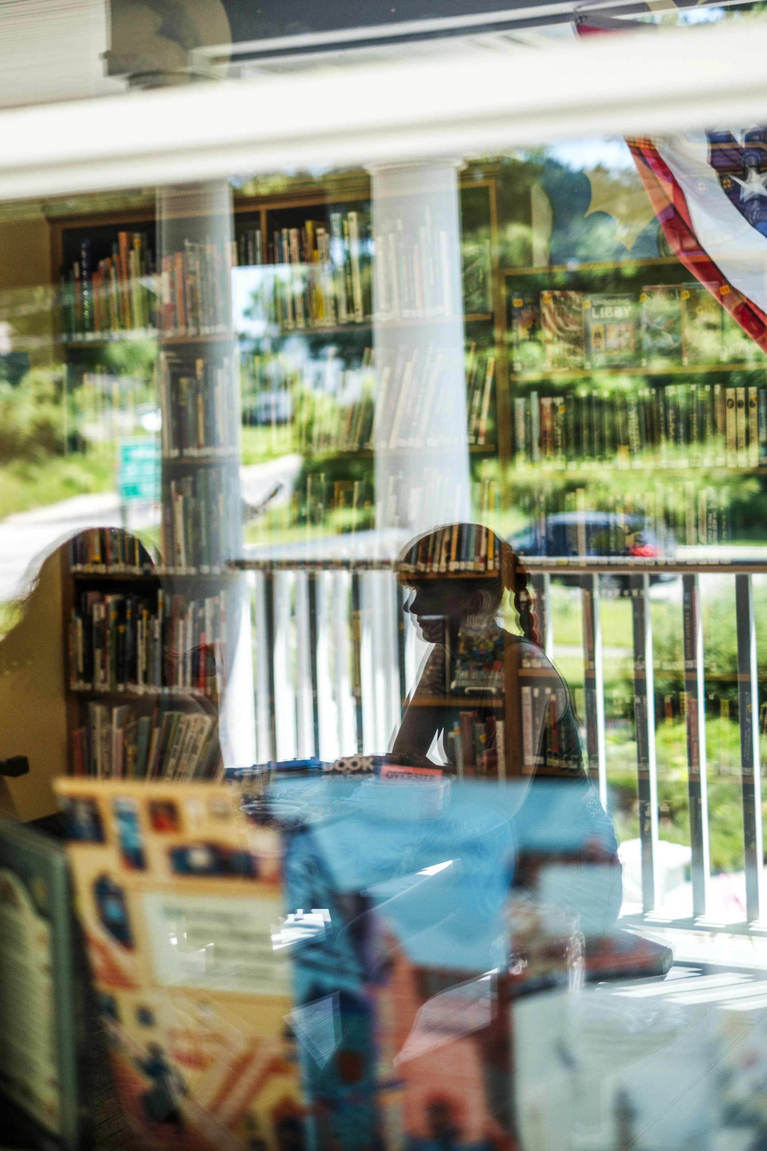 People are reflected in glass of a library, with shelves of books in the background.