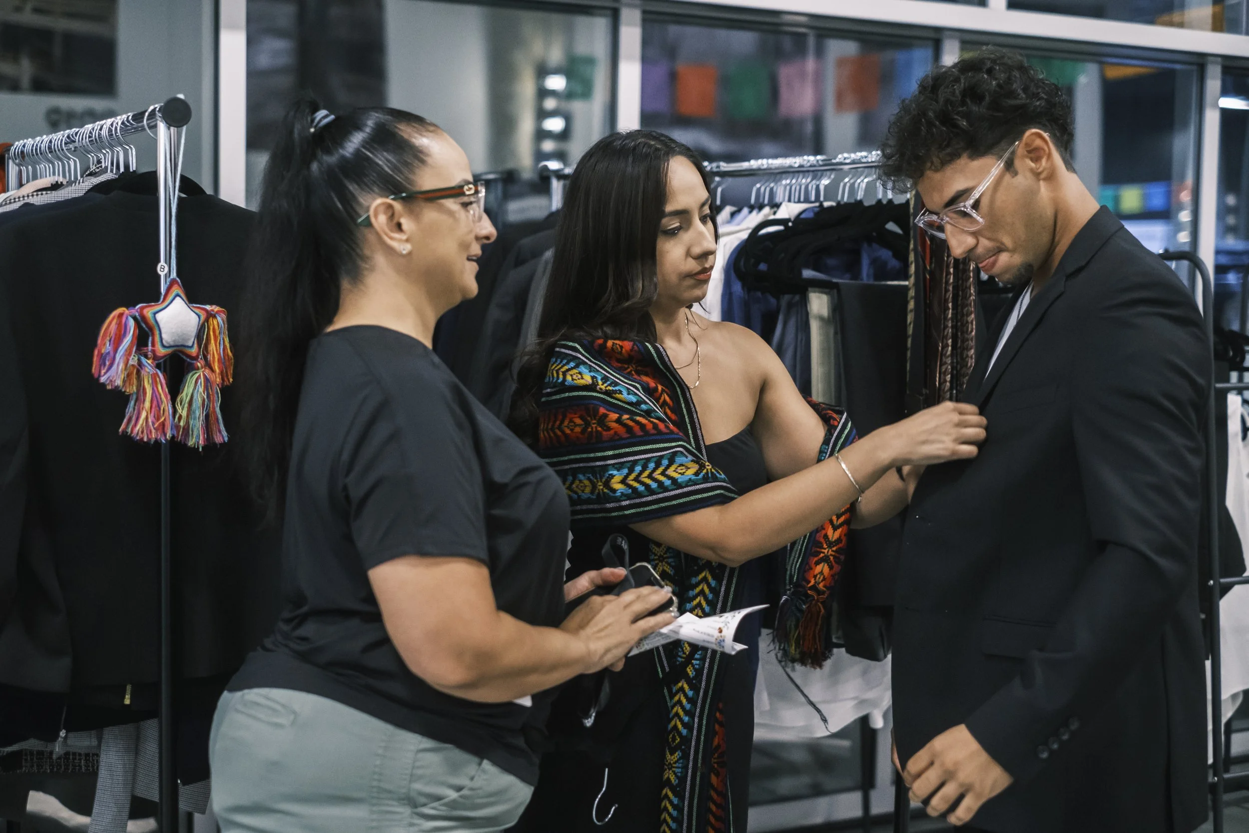Two women help a young man put on a suit jacket with clothing racks in the background.