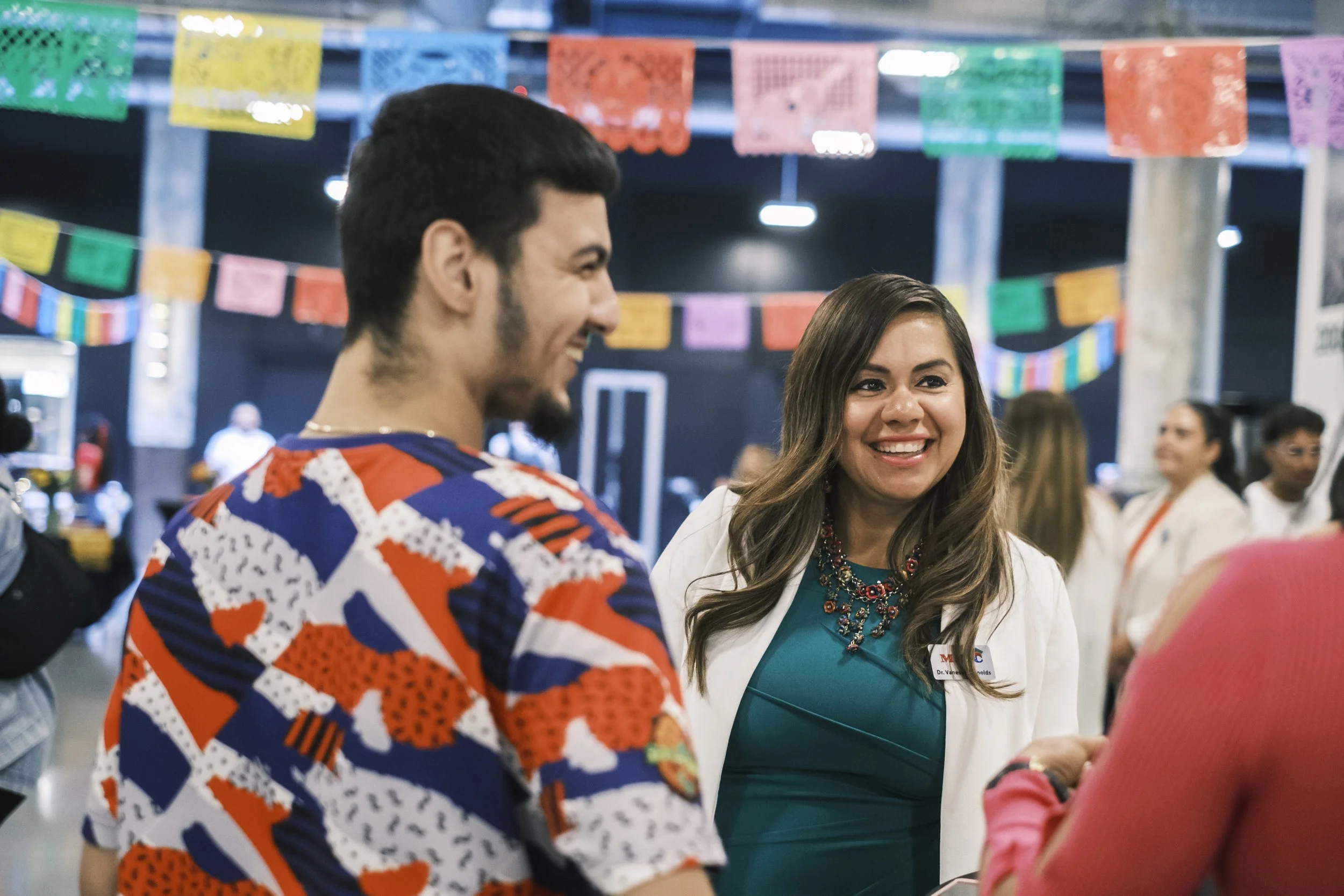 Two people talk with one another and smile with colorful hanging paper decor in the background.