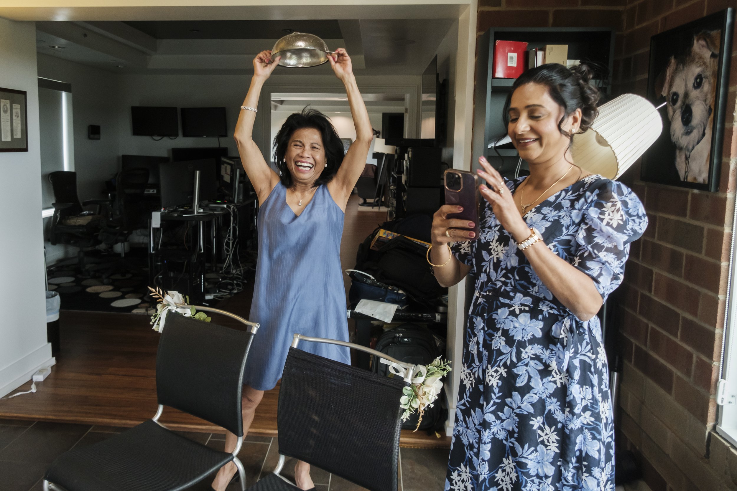 Two people, one holding a colander above her head and another holding her phone to take a picture in a room.