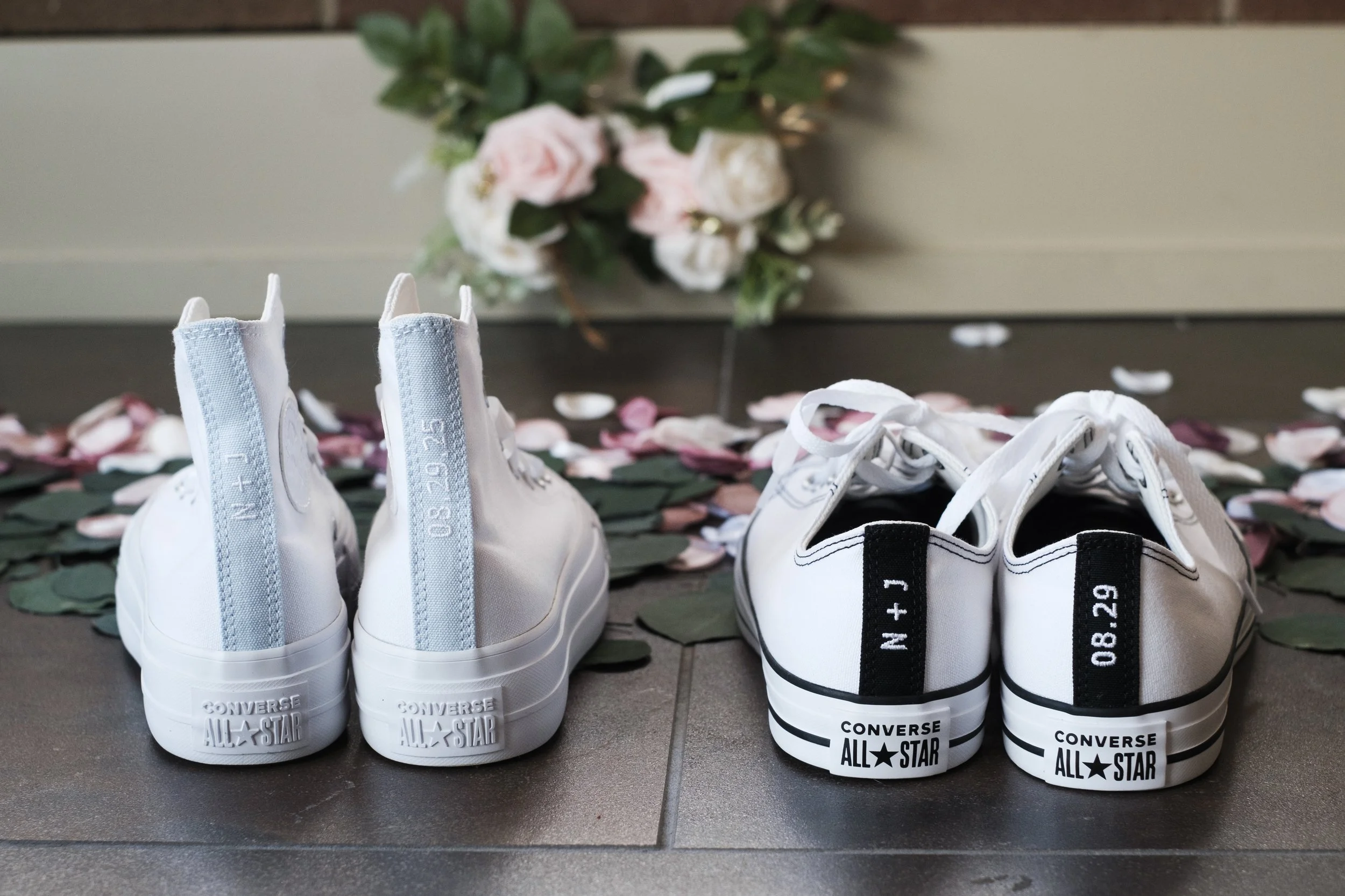 Two pairs of custom wedding converse sit on the ground with flowers in the background.