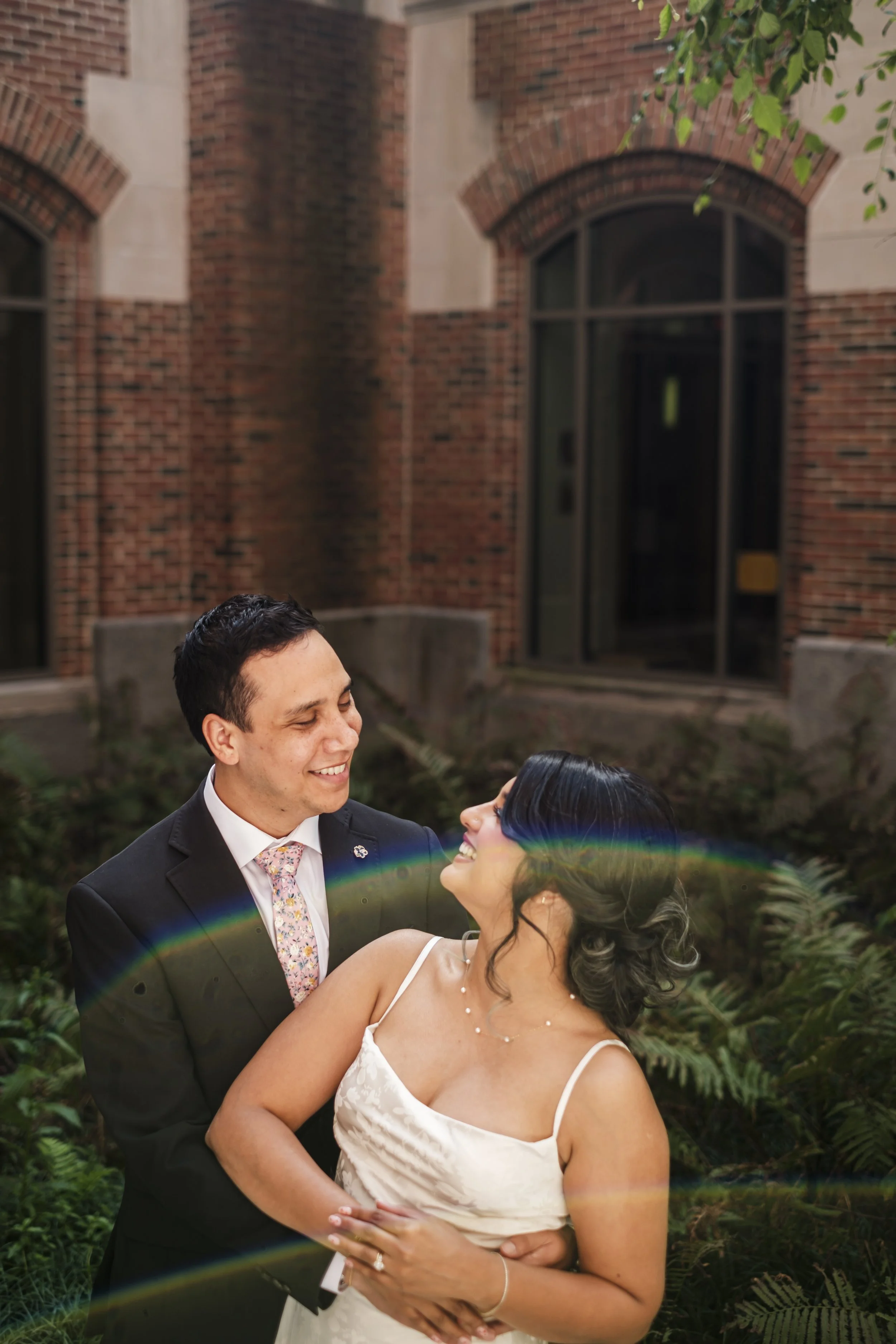 A bride and groom look at one another with a light rainbow flare in the foreground and brick building in the background.