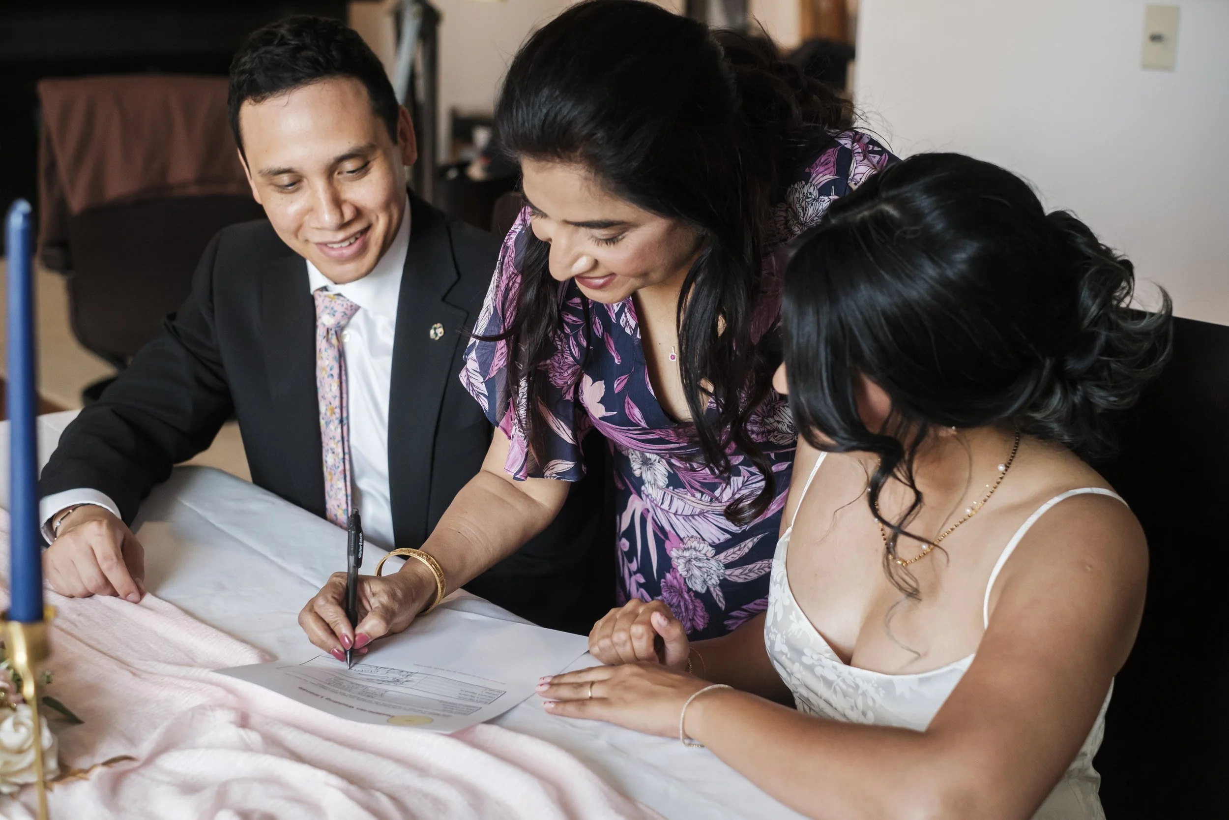 Three people sit at a table and sign a document during a wedding.