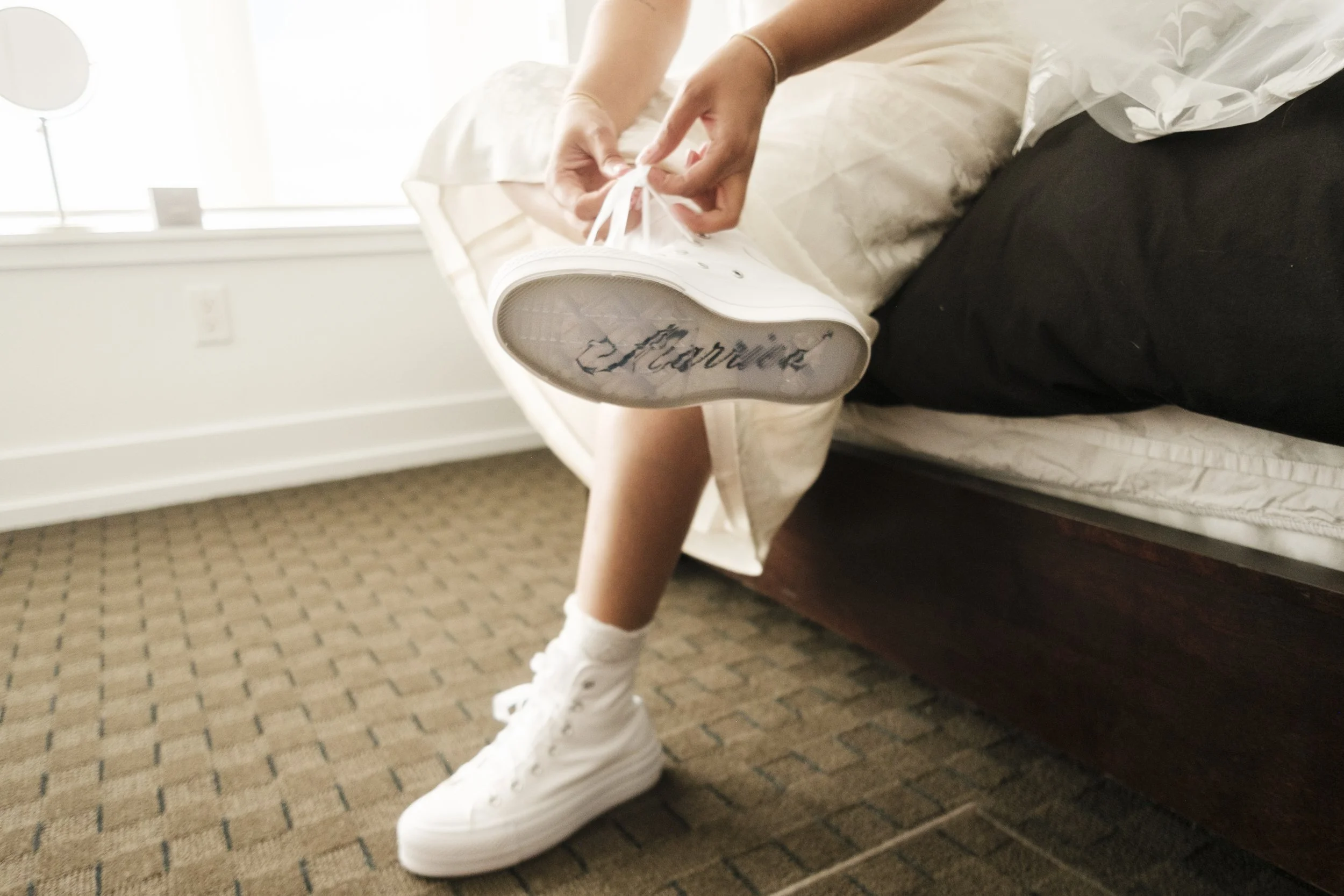 A bride ties her custom converse ahead of her wedding. The bottom of the converse say the word "bride".