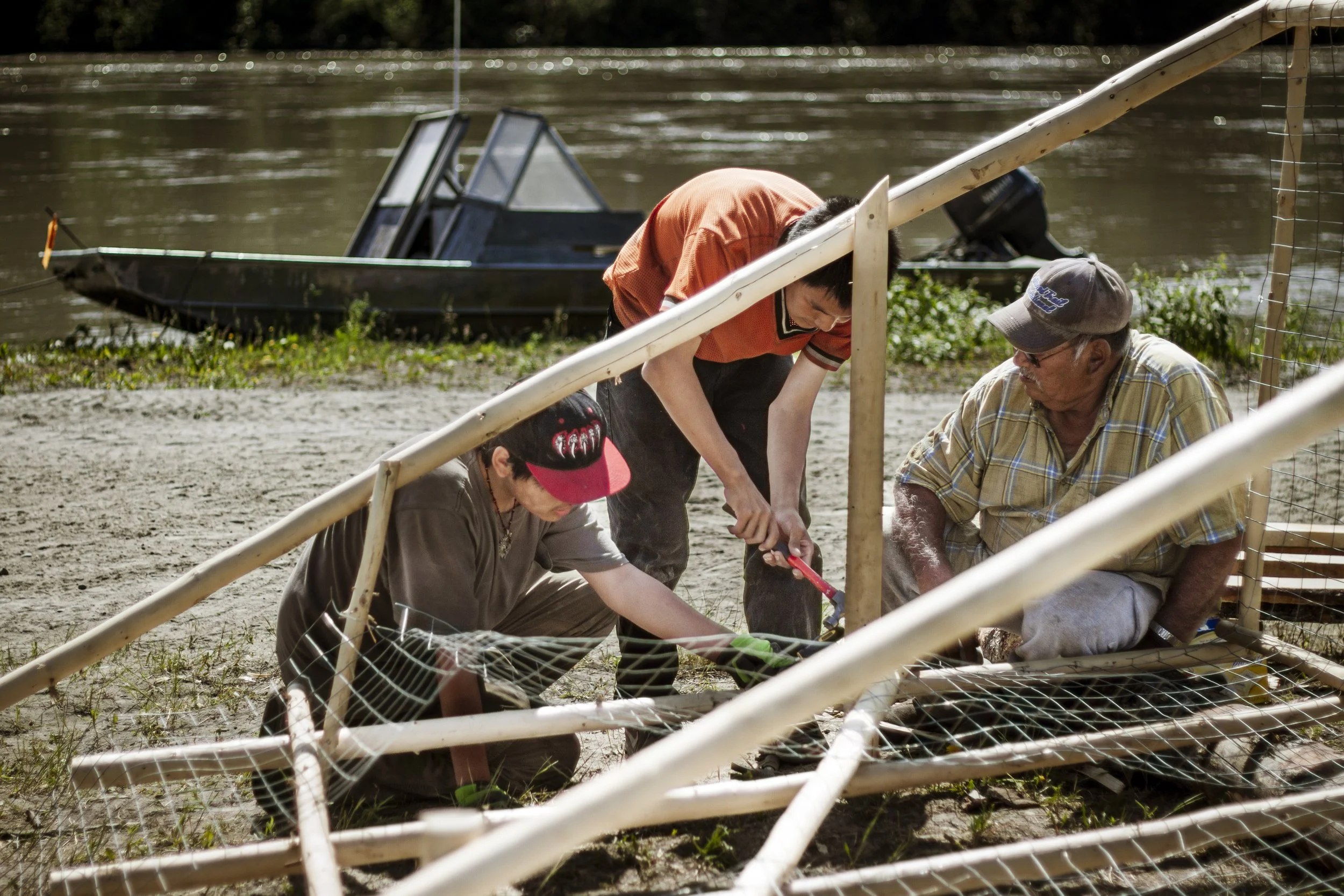 Three people, two young men and an elder, sit around wood posts and use a tool with a river in the background.