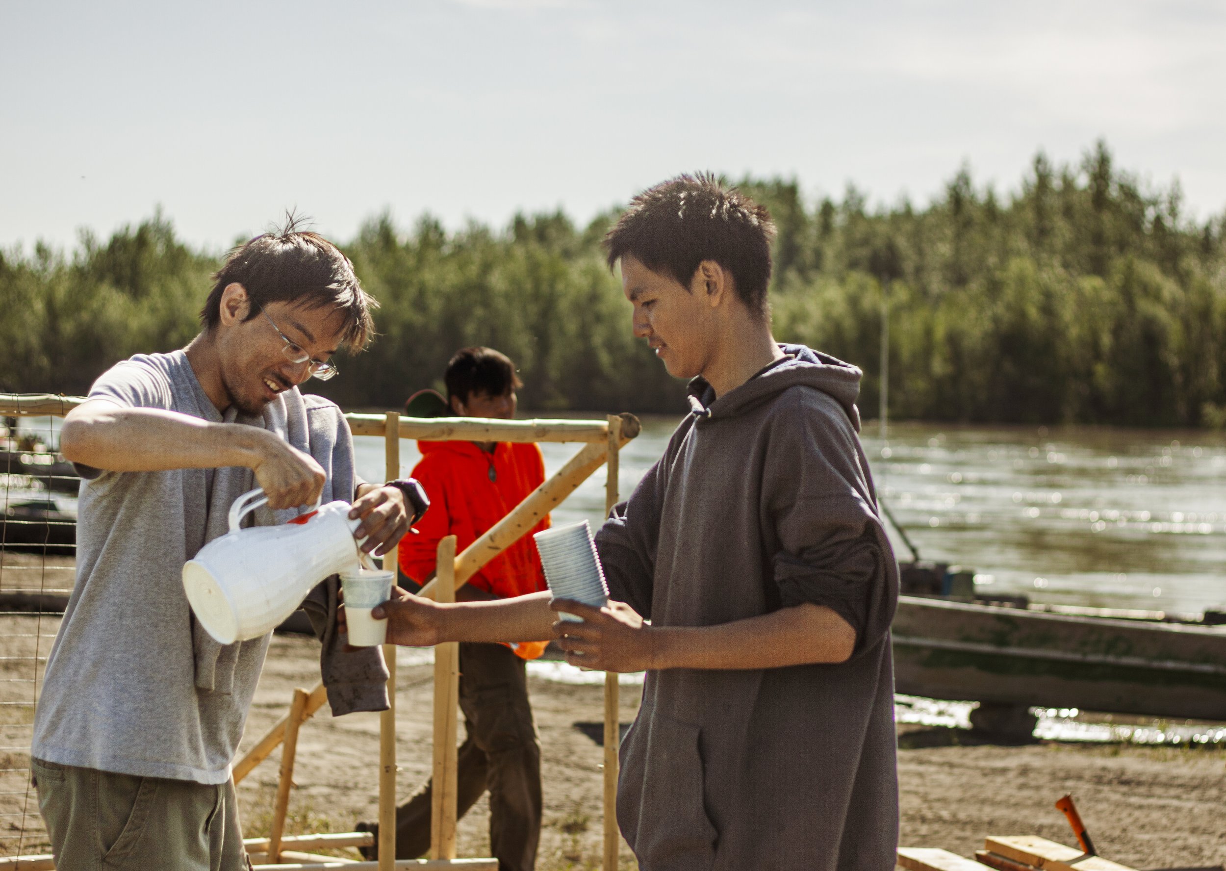 Two people stand in the foreground, one holding coffee cups and another pouring tea from a teapot with trees the Kuskokwim river in the background.