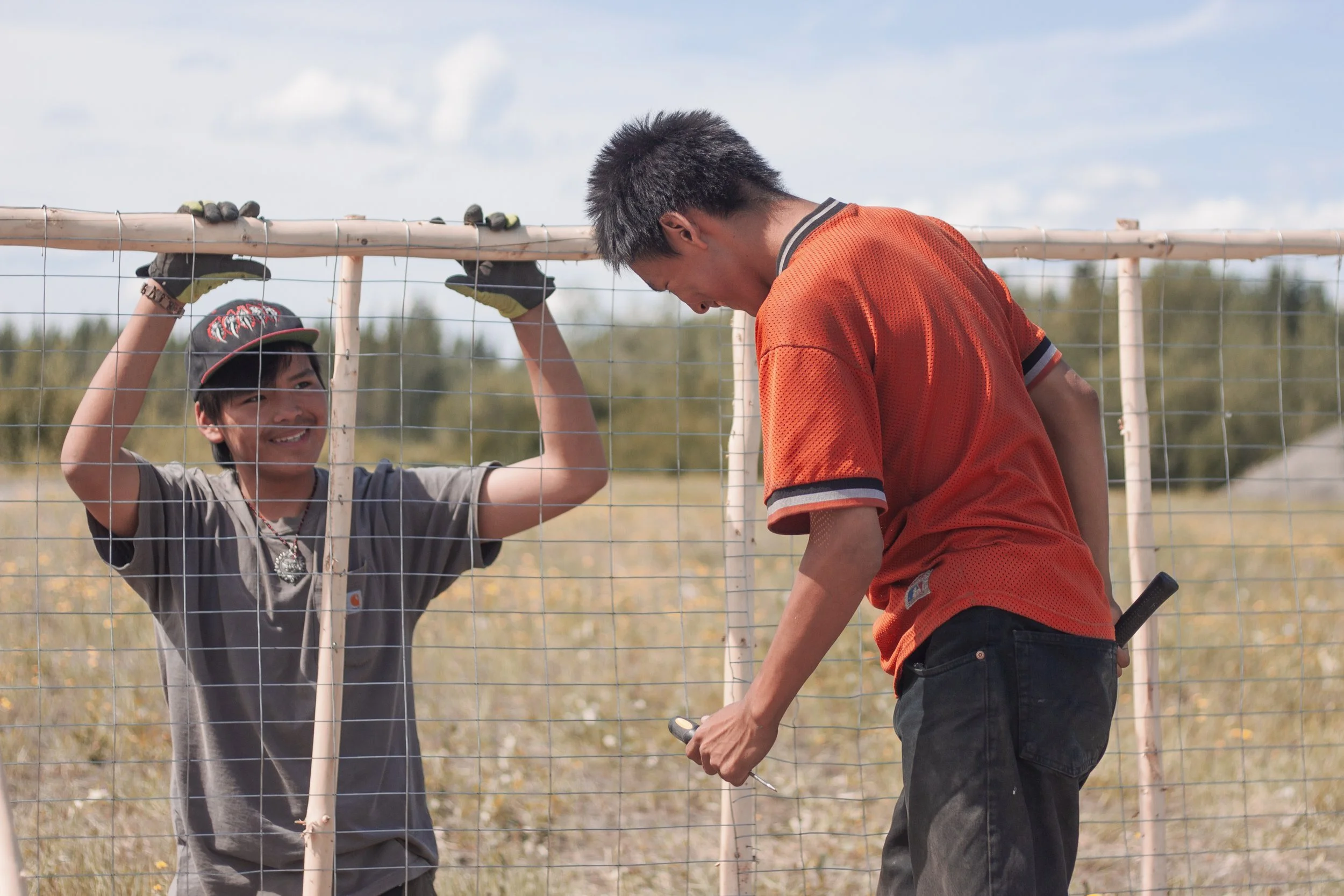 Two young men, smile at one another while holding tools while working on a wooden fishwheel.