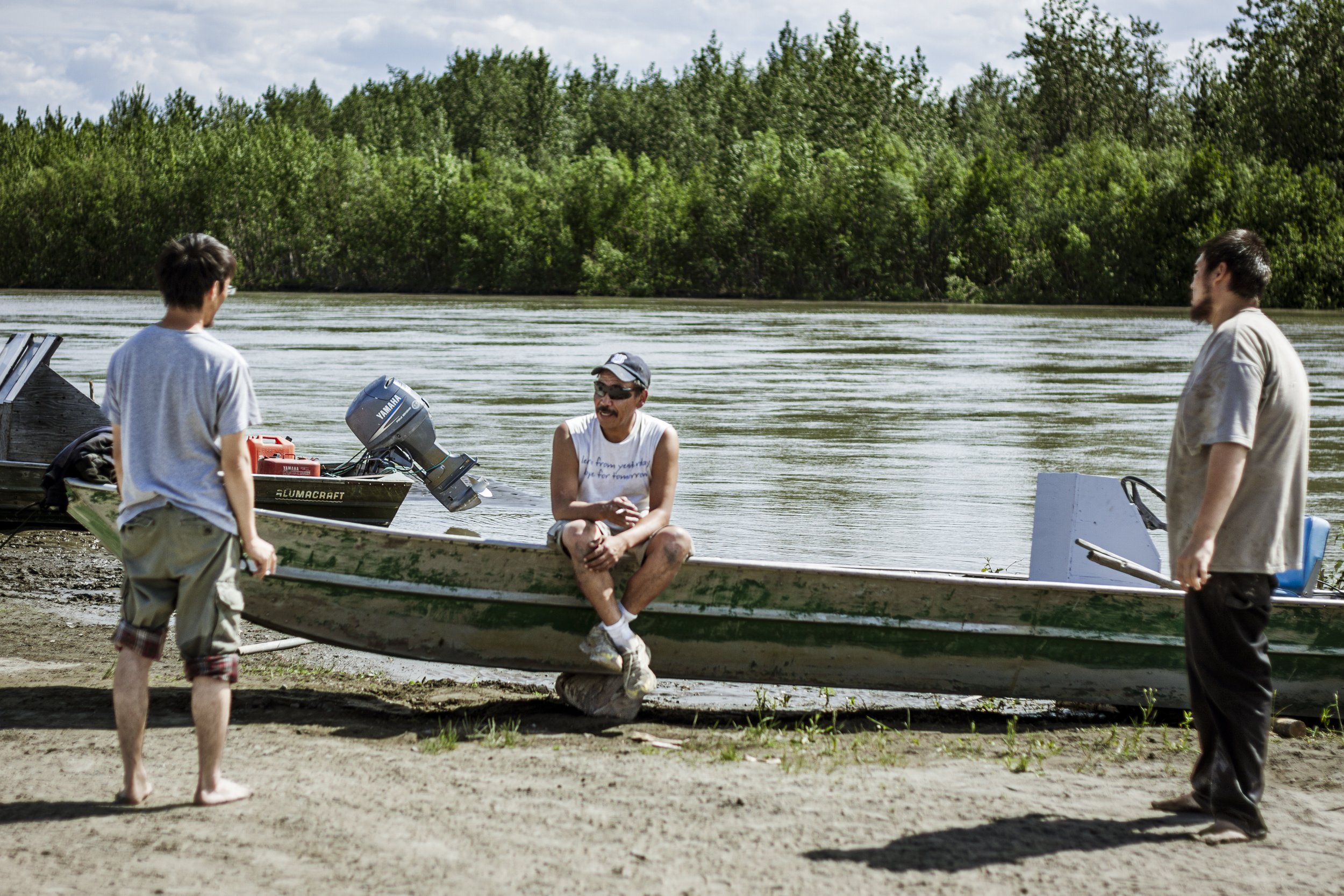 Three men, two standing and one sitting on a fishing boat, talk with one another, with the Kuskokwim river and remote Alaskan wilderness in the background.