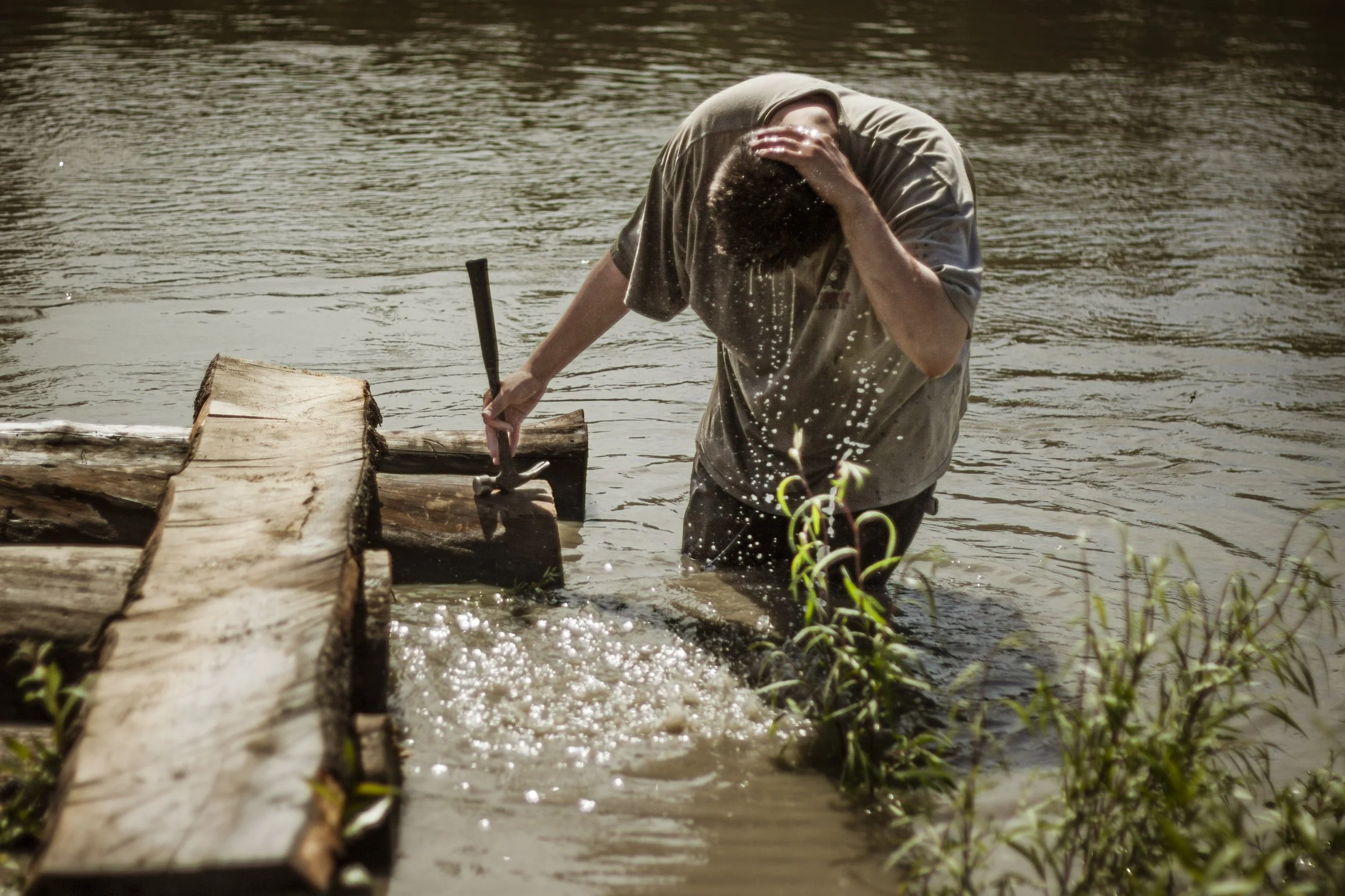 A man uses his hand to cup river water that he is standing in to pour it over his head while holding a hammer in his other hand.