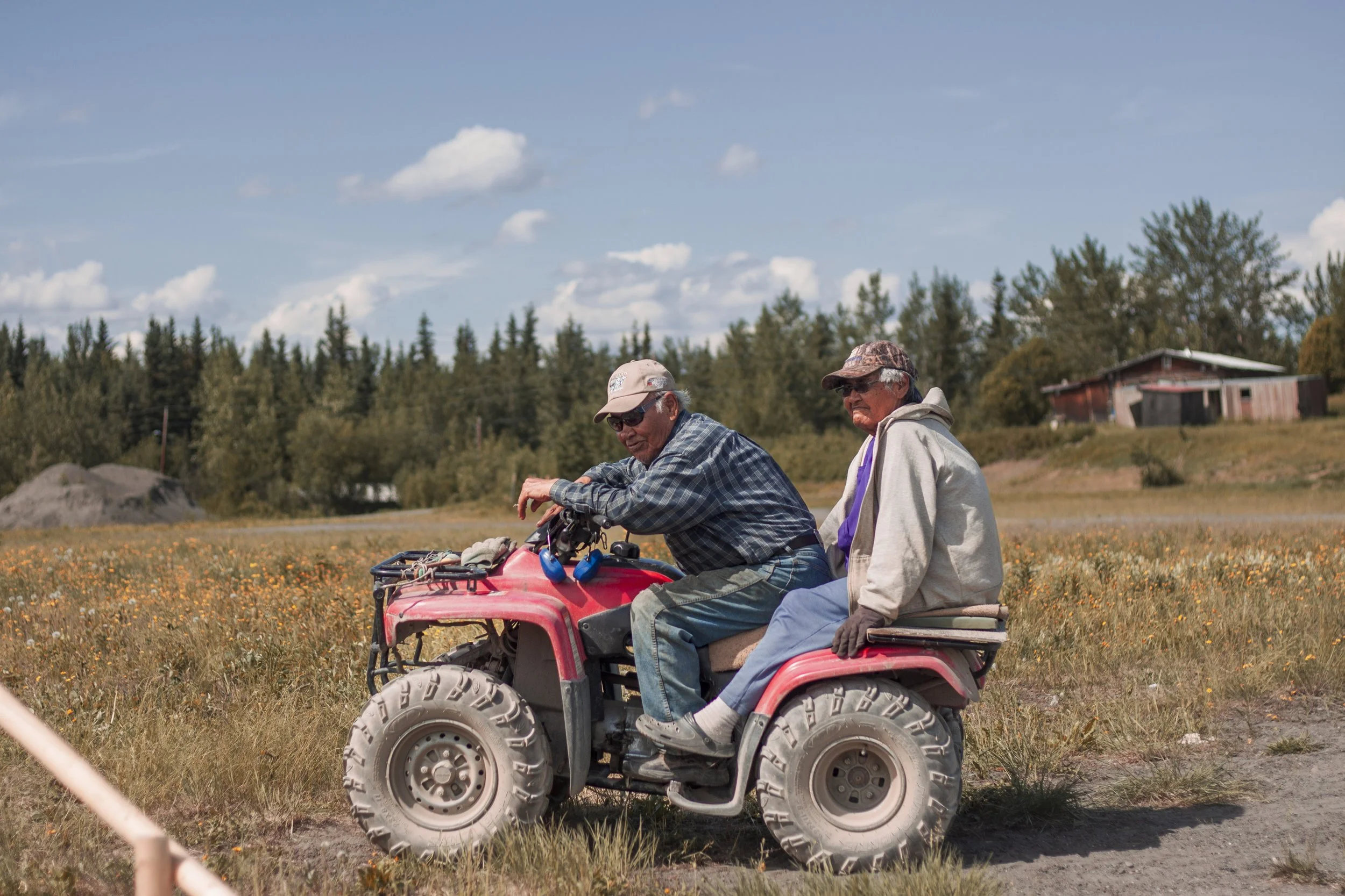 Two elders, one man and one woman, sit on a four wheeler under a blue sky with trees and a house in the background.