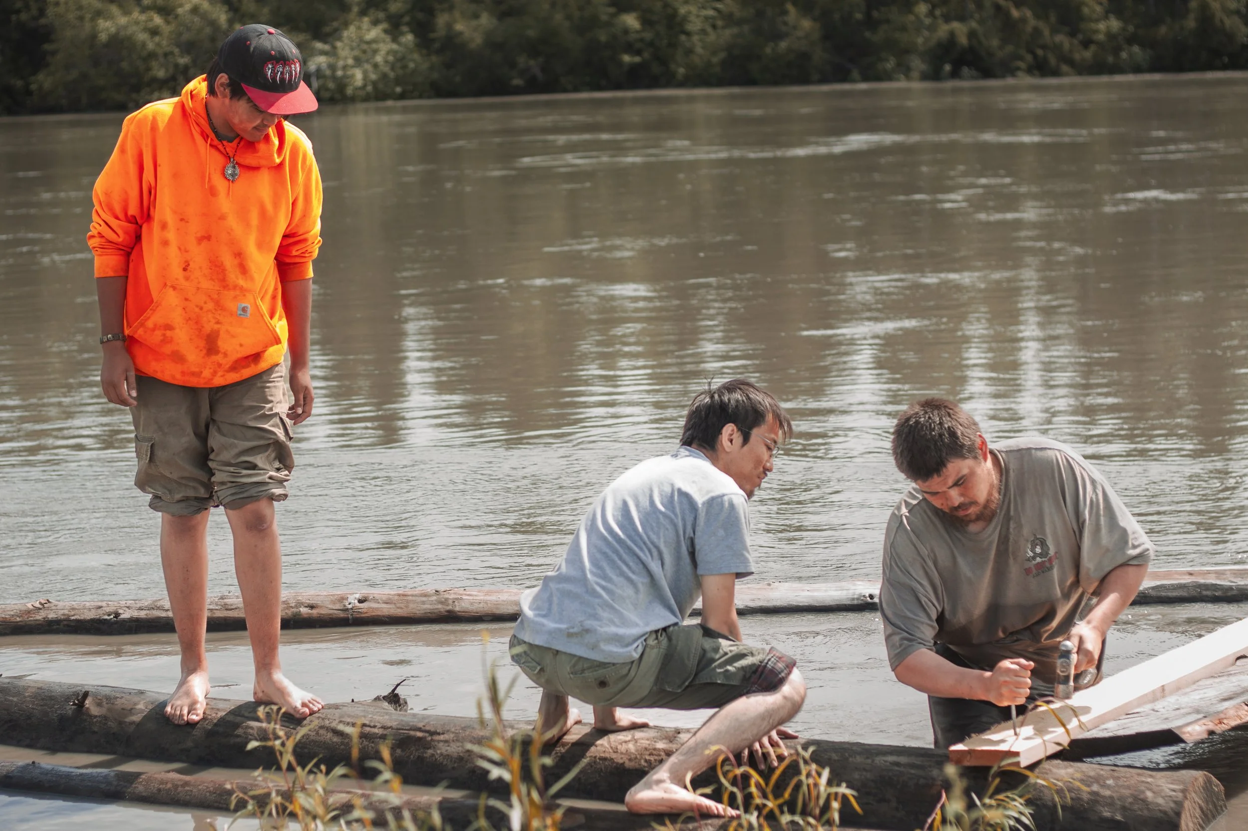 A young man stands barefoot on a log, wearing a bright orange sweatshirt, watches two other young men nail a wooden board onto a log while standing in a river.