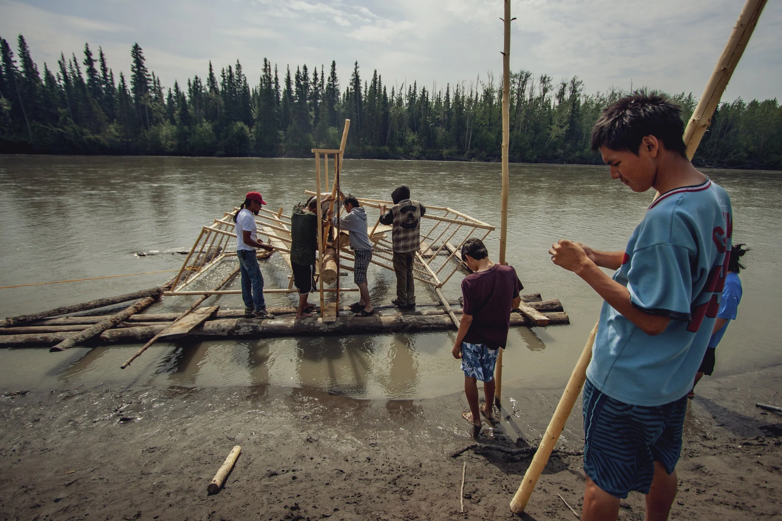 A wide perspective showing the alaskan wilderness while a group people work on a fishwheel. Four men work together to assemble logs onto the fish wheel while standing in a river, while two young men wait next to them, holding wooden posts.