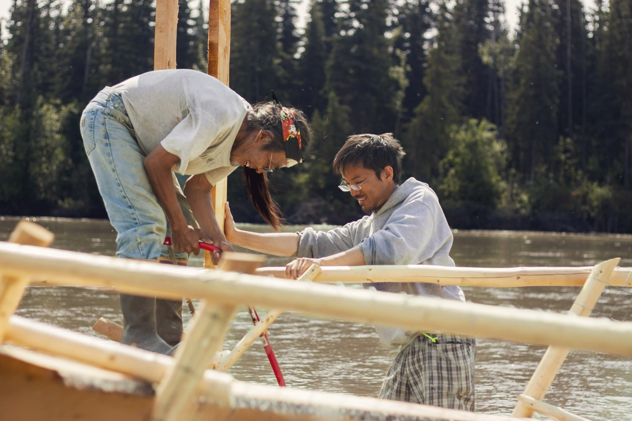 Two men smile with one another while holding wooden posts together to construct a fish wheel, while standing in a river with trees in the background.