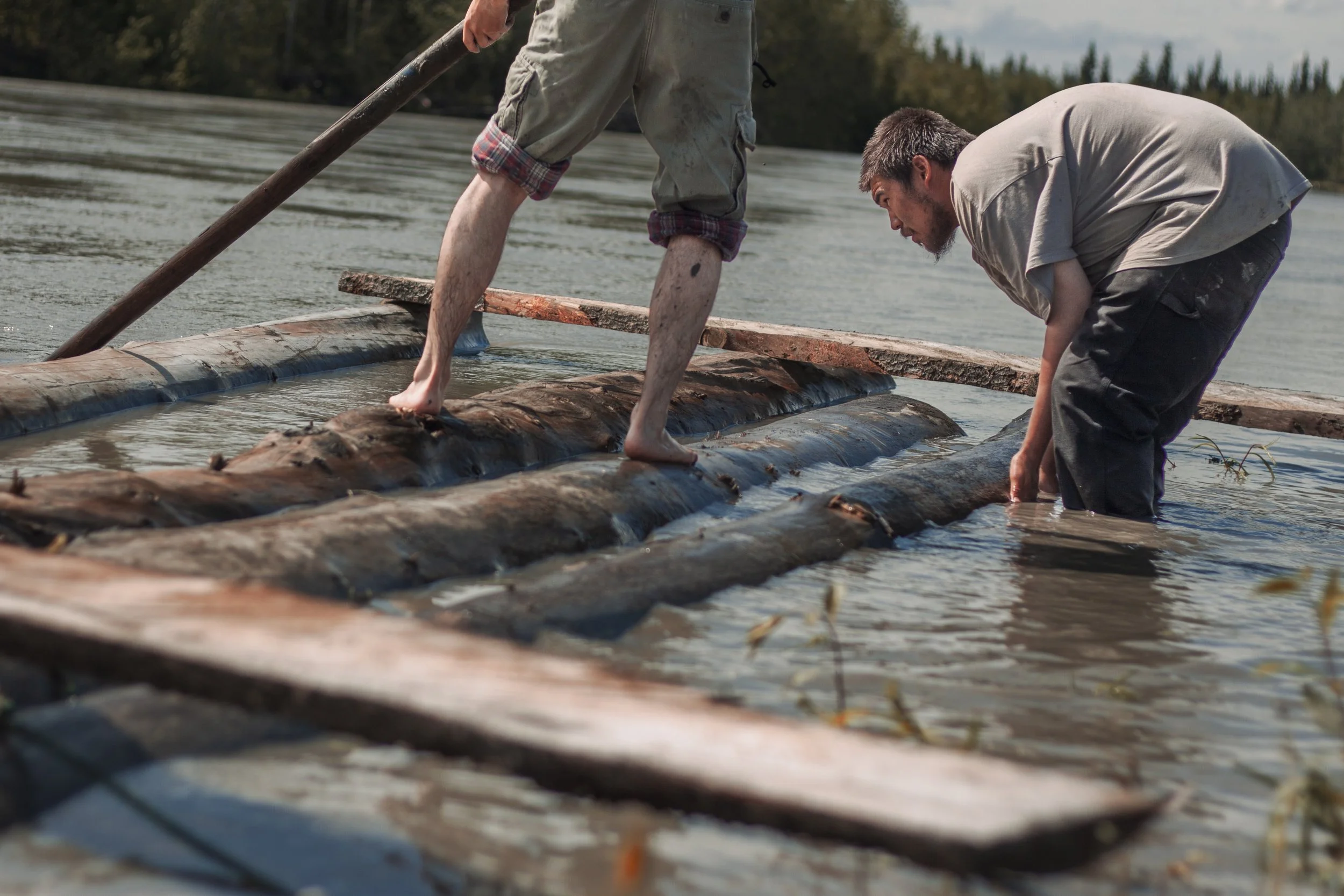 A tilted perspective with feet walking on logs and a man pushes logs together while standing in a river.