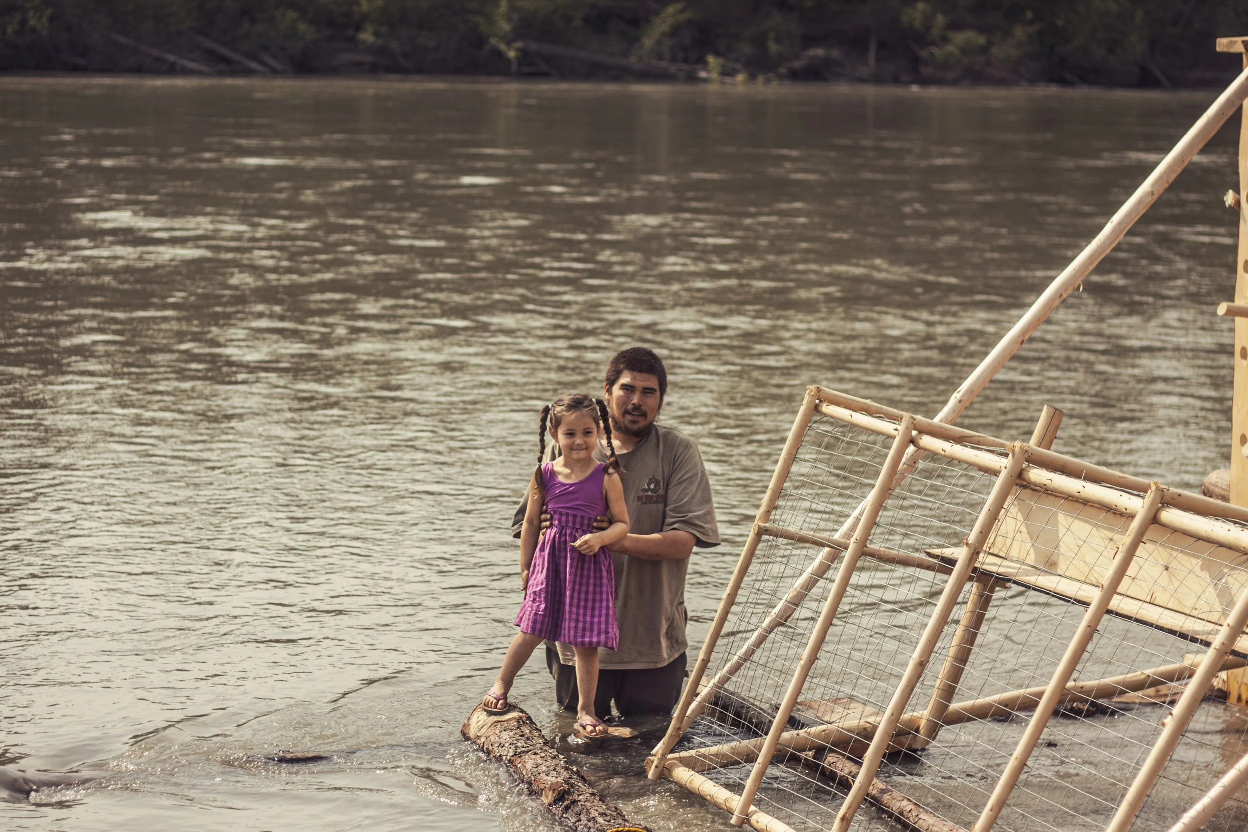 A young girl smiles in a bright purple dress stands on logs in a river while being held by a young man, who holds her up.