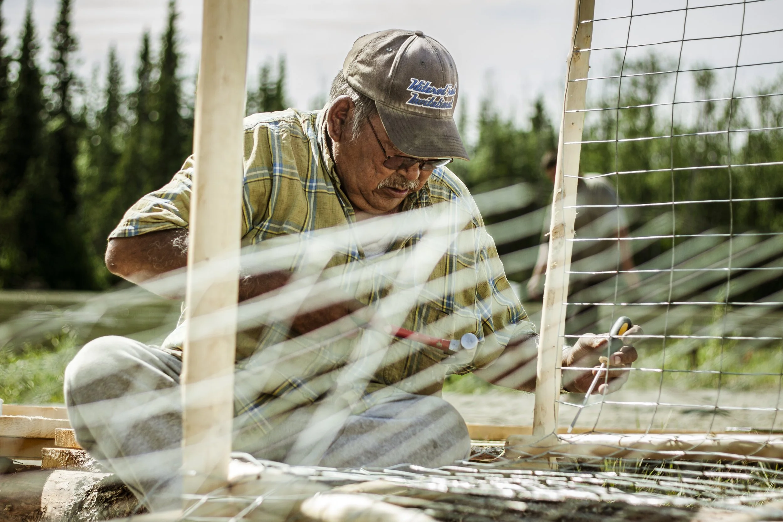 An elder wearing a baseball and green plaid shirt sits cross legged while holding a hammer in one hand and uses his other hand to wrap metal netting around a pole.