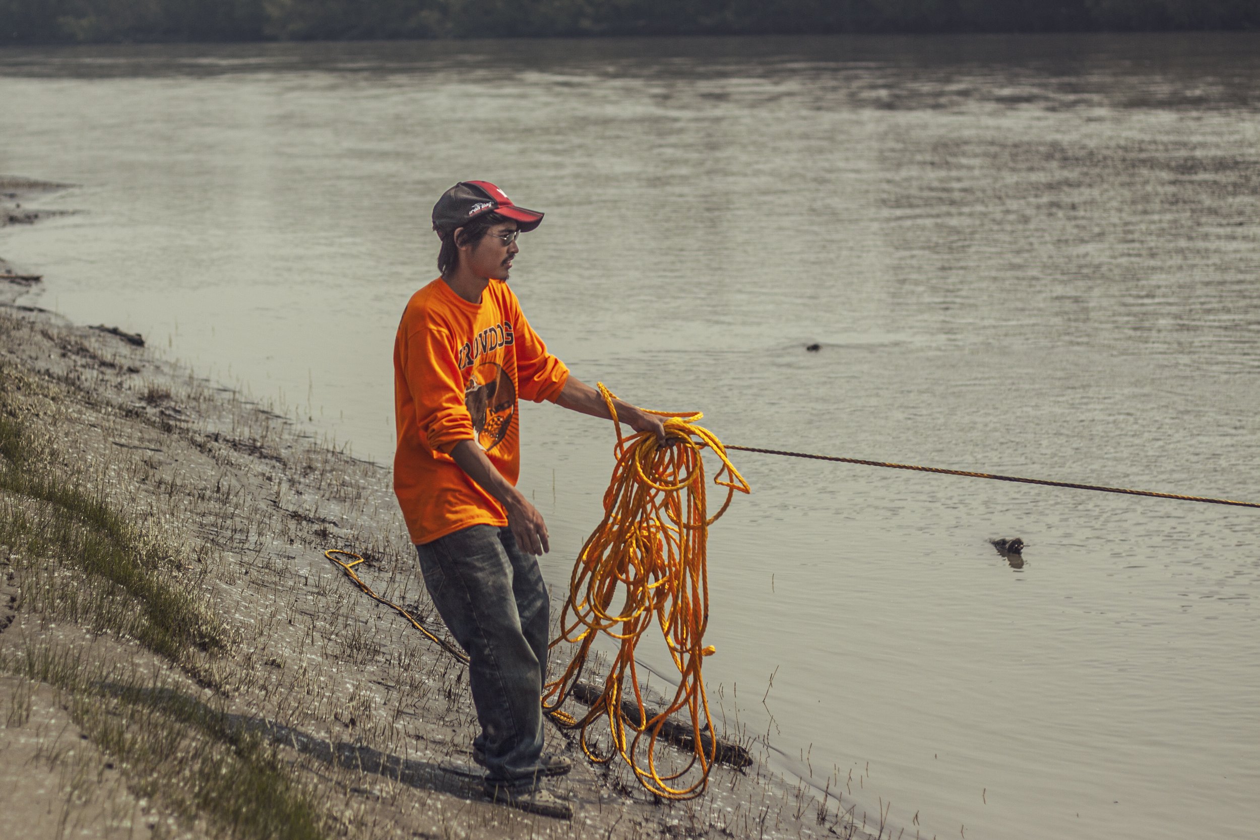 A young man stands on the muddy shoreline of the Kuskokwim river while holding lots of rope.