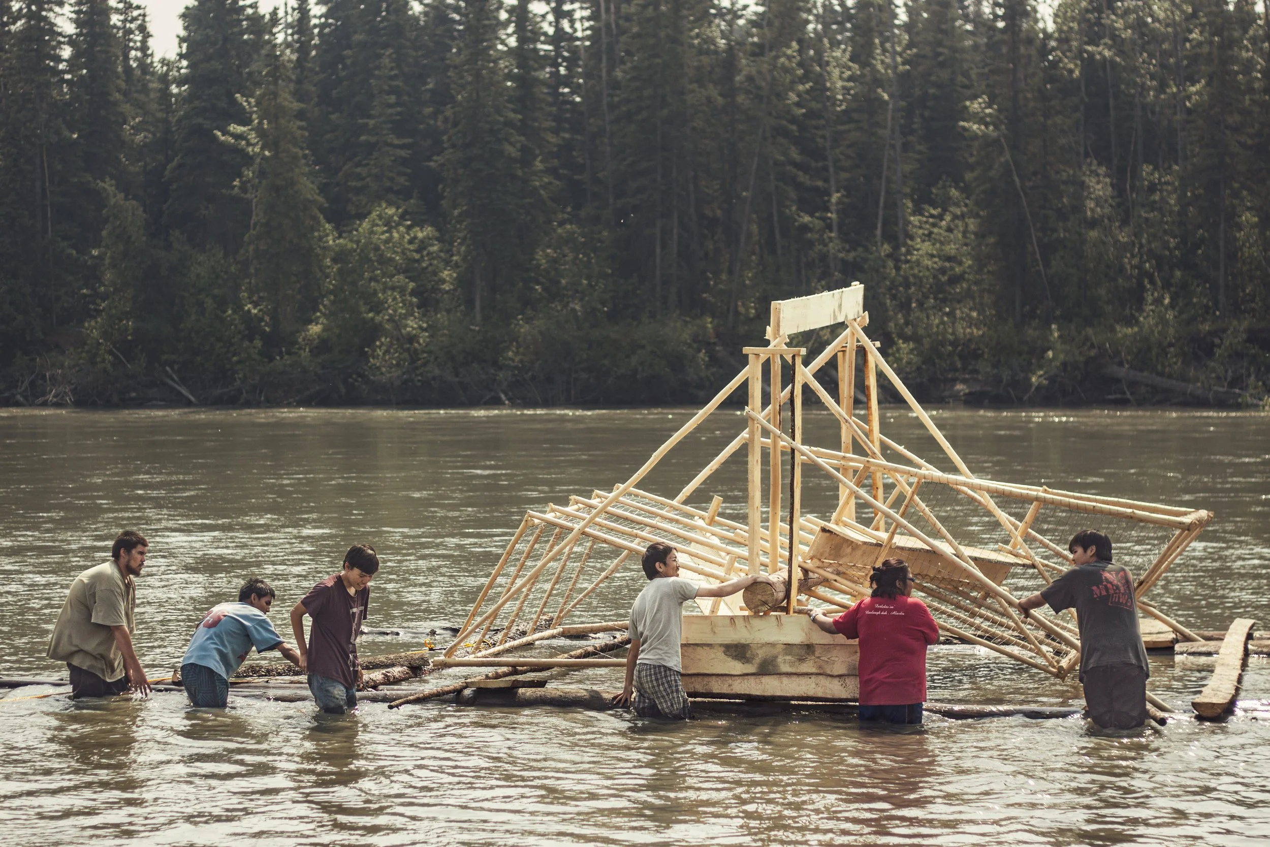 A group of young men and a young woman stand in a river while holding a fish wheel, a large wooden device with baskets that is used to catch fish.