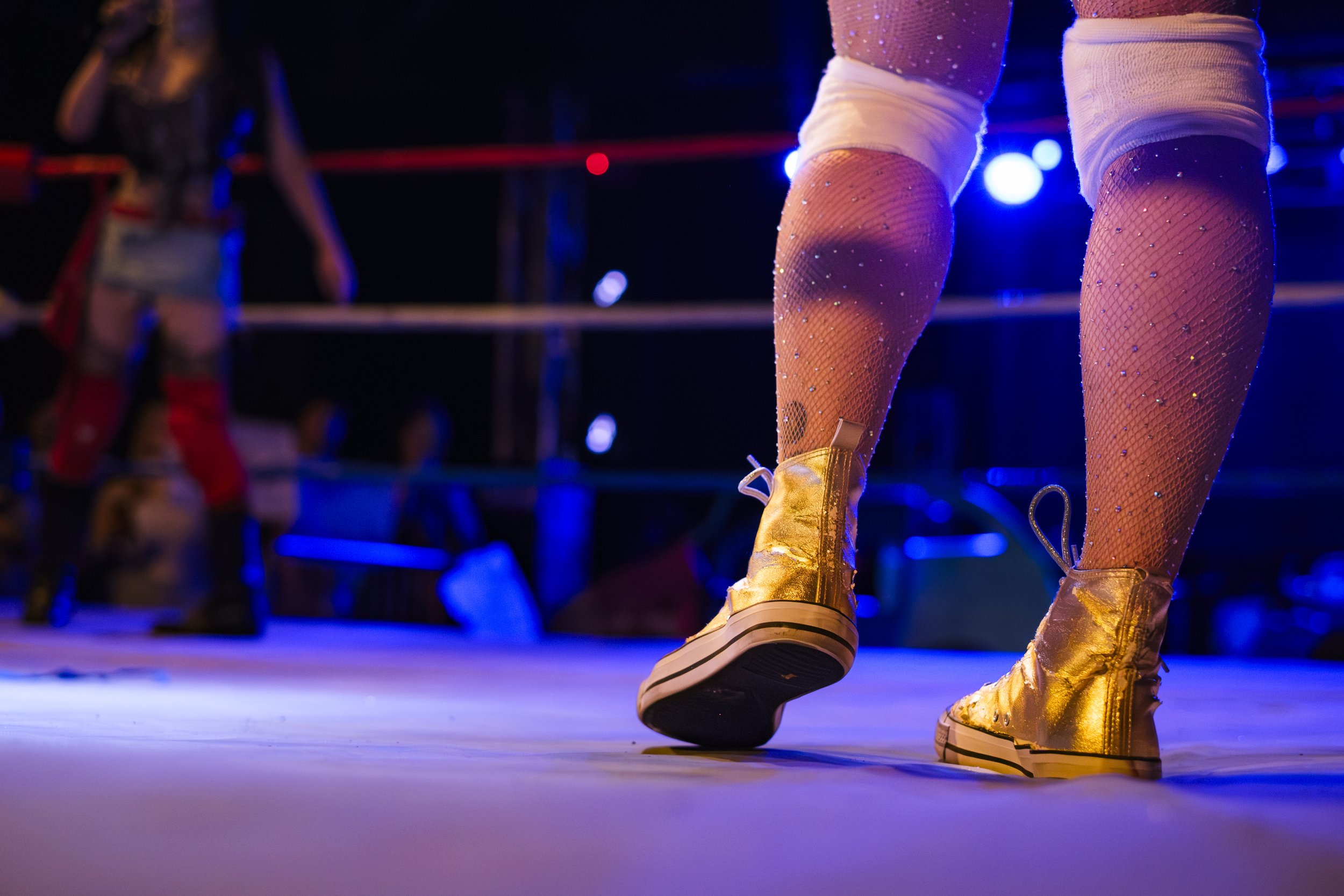 Gold converse shoes and pantyhose with sparkles in a wrestling ring, illuminated by blue light.