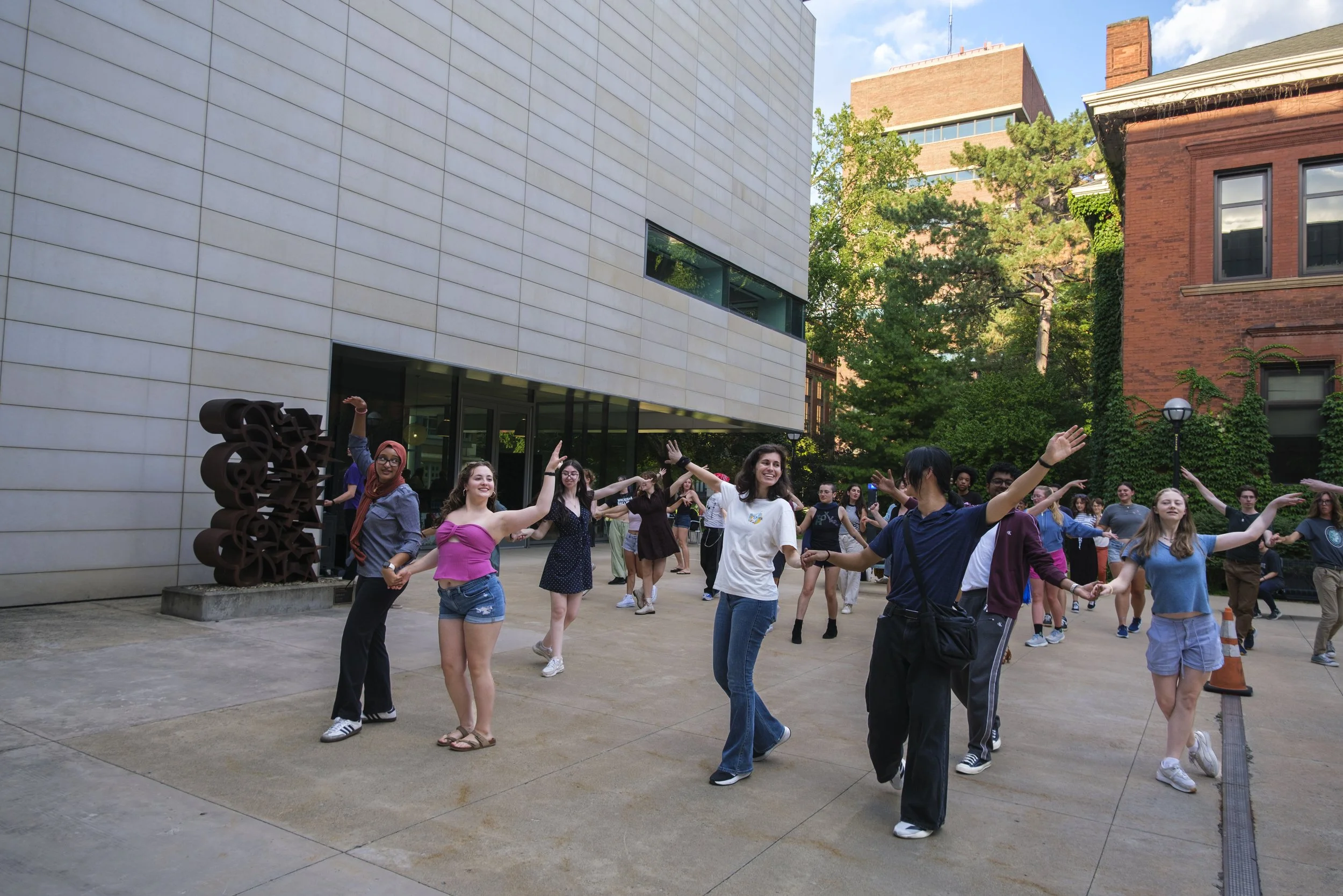 A group of people participate in swing dancing lessons in the side yard of UMMA, outdoors while smiling.