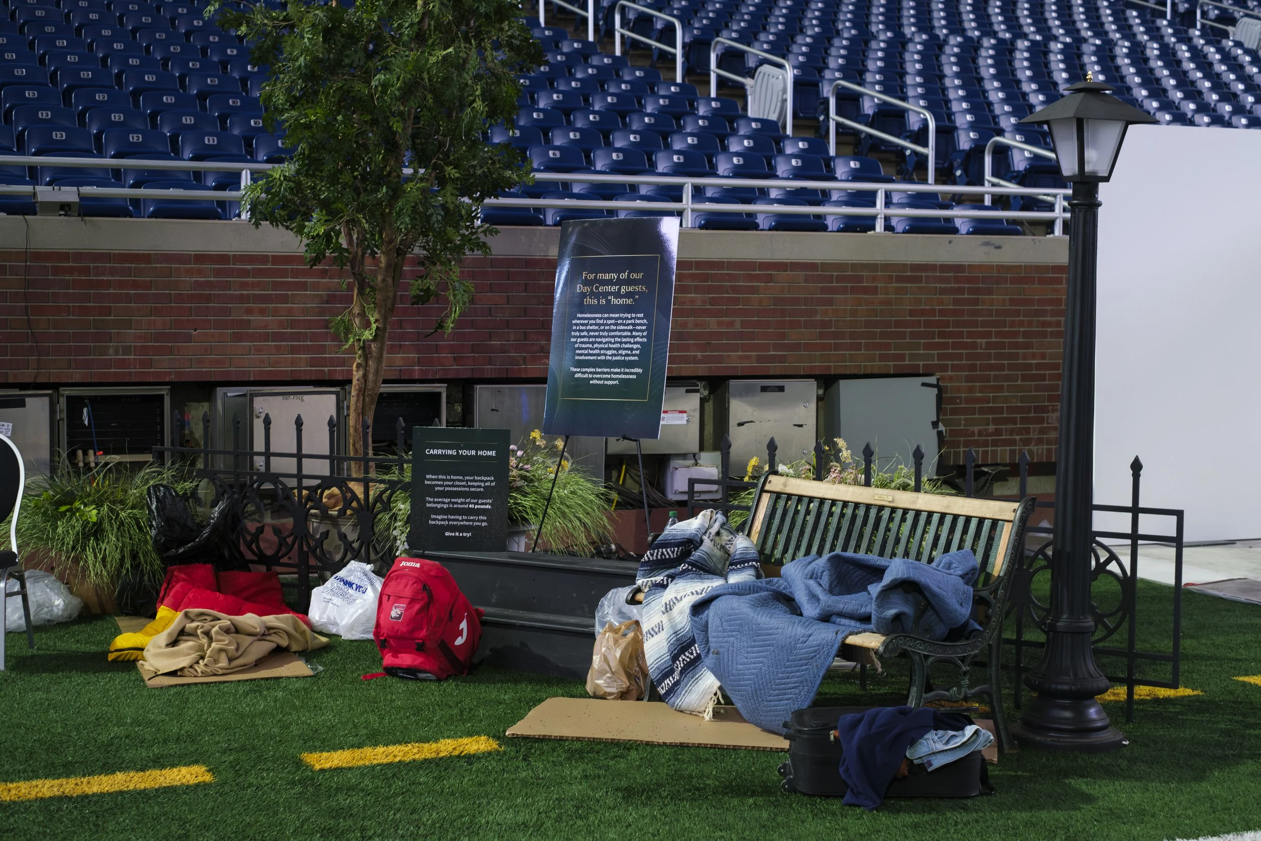 A display featuring a backpack, blankets, a bench and light post are set up on turf. A sign next to the items reads "For many of our Day Center guests, this is home".