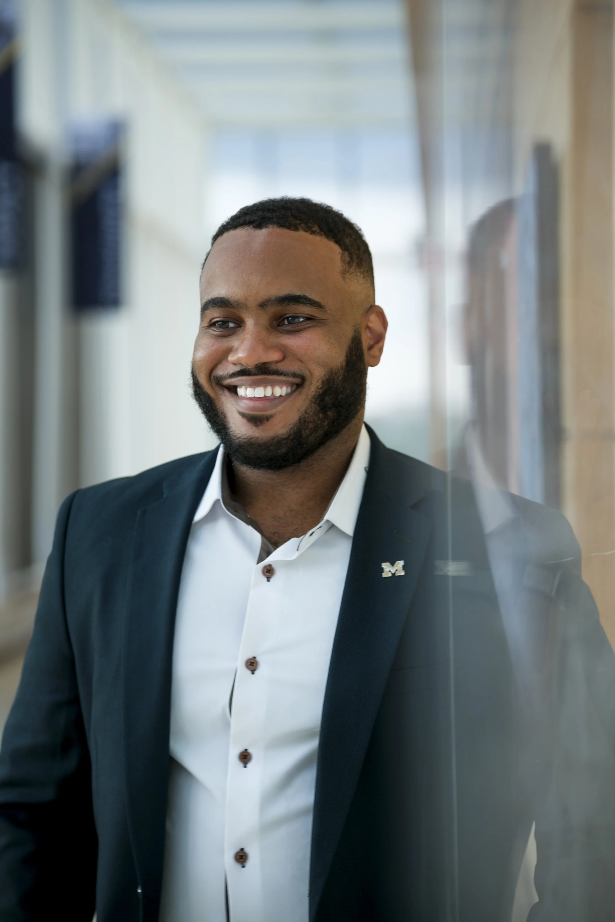 A man wearing a suit with a Block M pin on his lapel smiles.
