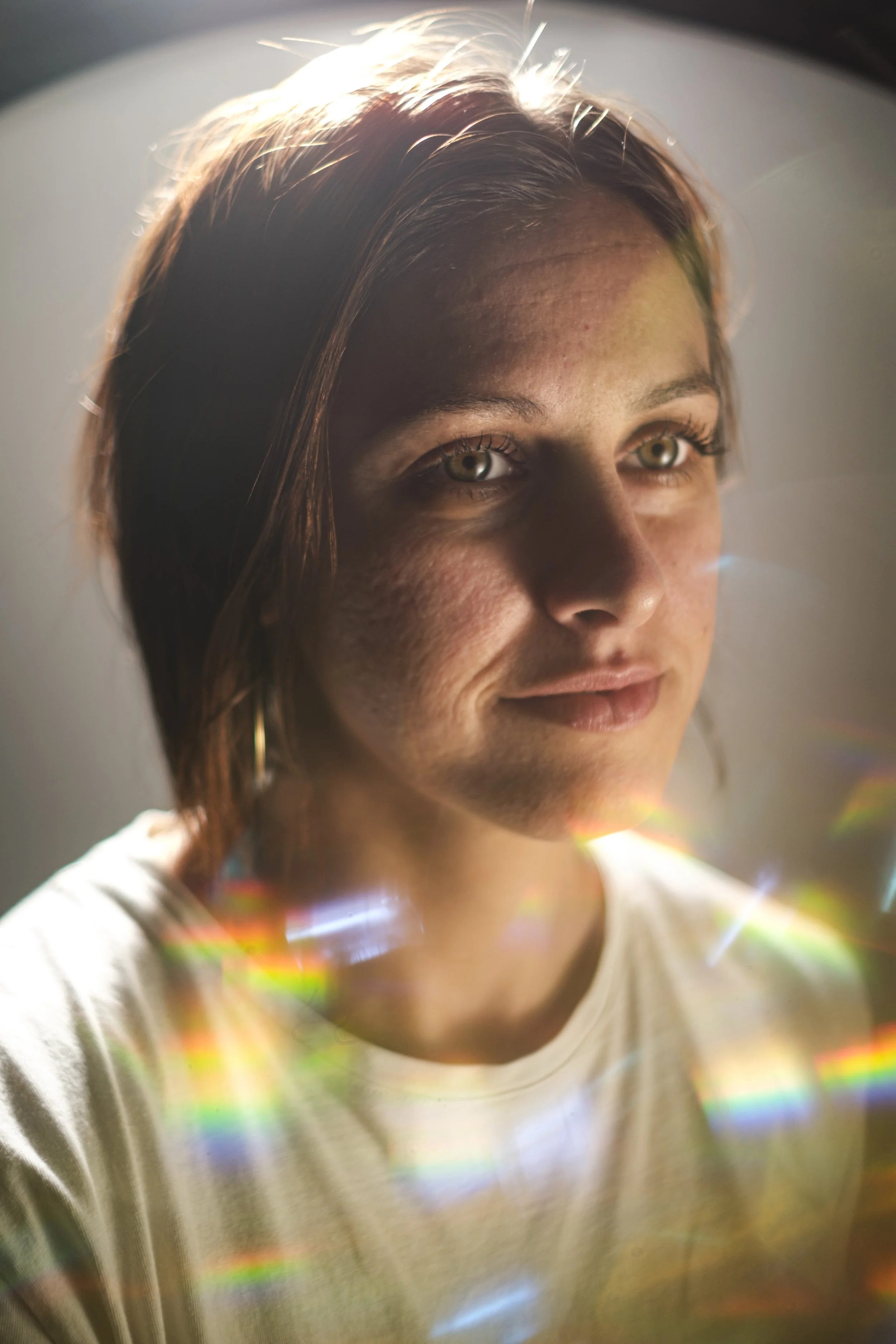 A photo of a young woman sitting against a gray backdrop with rainbows in the foreground.