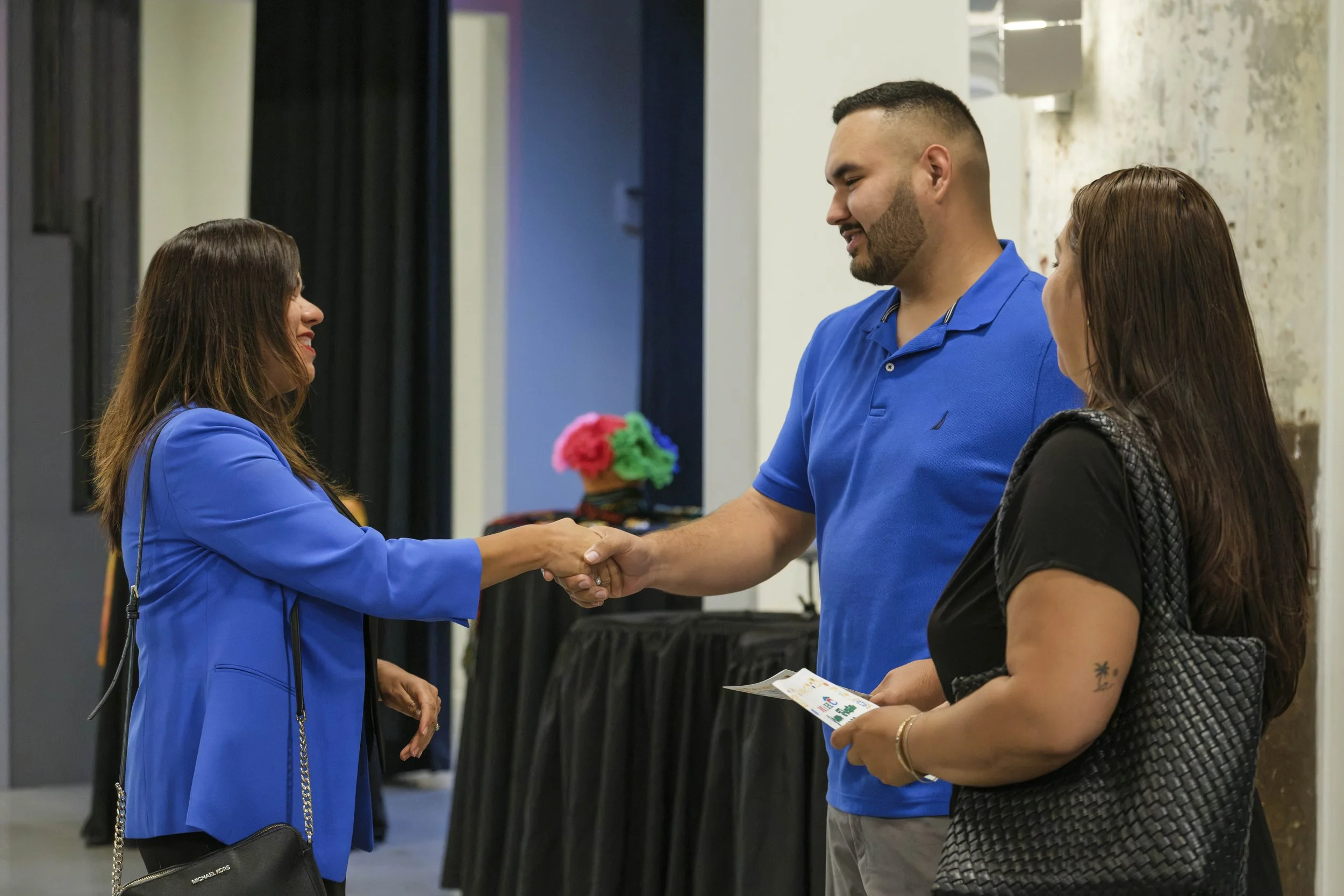 A woman in a blue blazer shakes the hand of a young man in a blue polo shirt with black high top tables in the background.