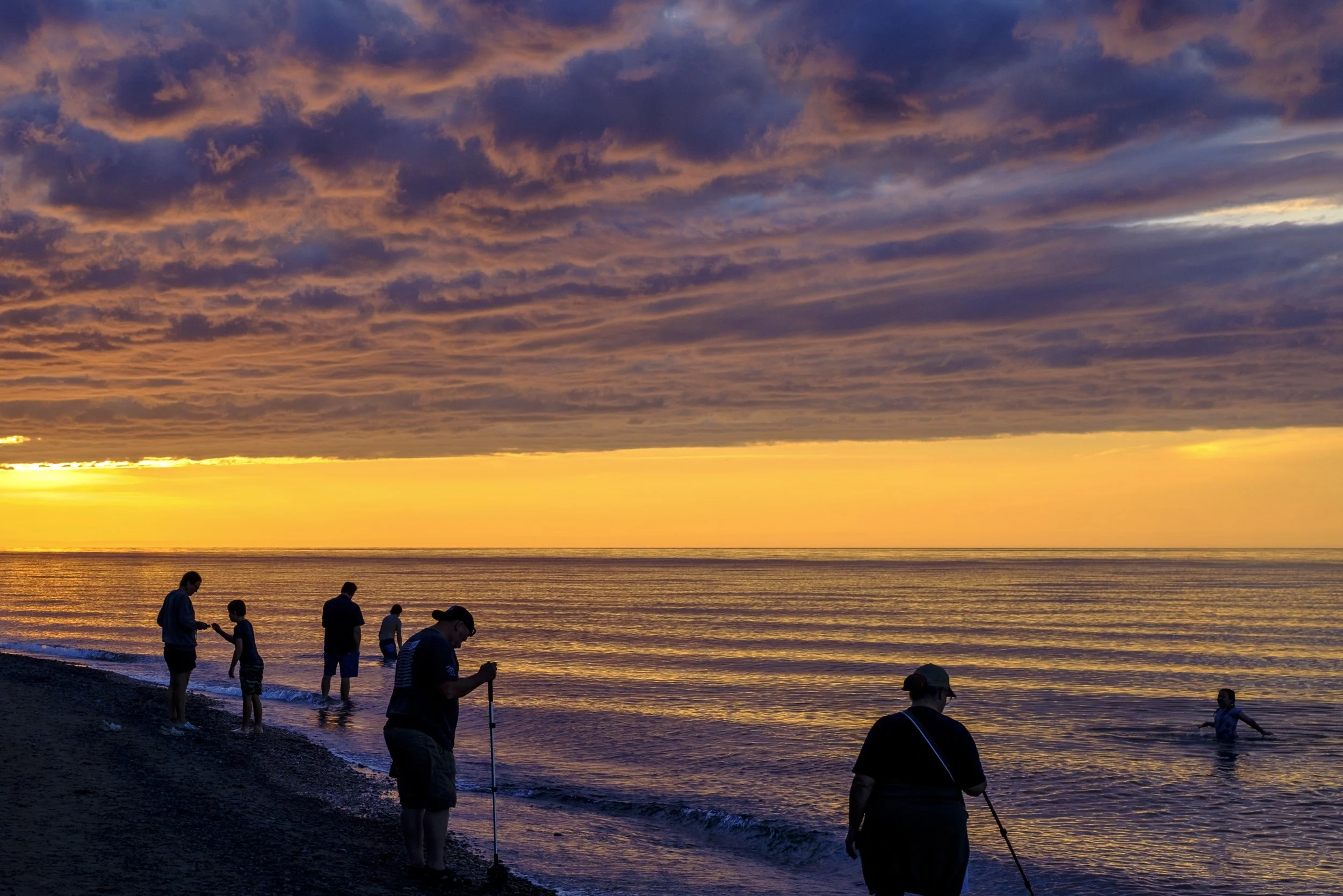 A gold and purple sunset outlines people using rock hunting tools on the shores of Lake Superior in Michigan's Upper Peninsula.
