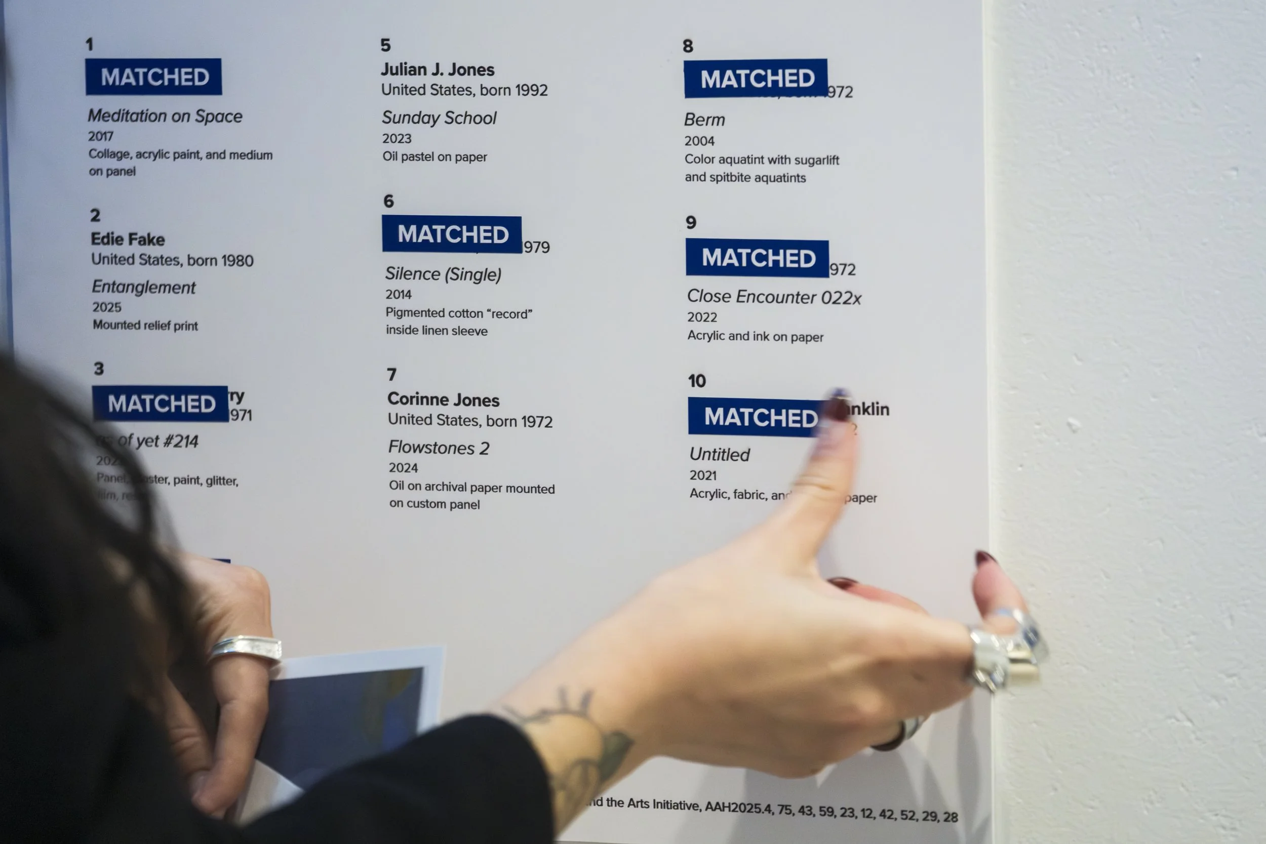 A hand reaches out to put a sticker that says "matched" on a board with names of artwork.