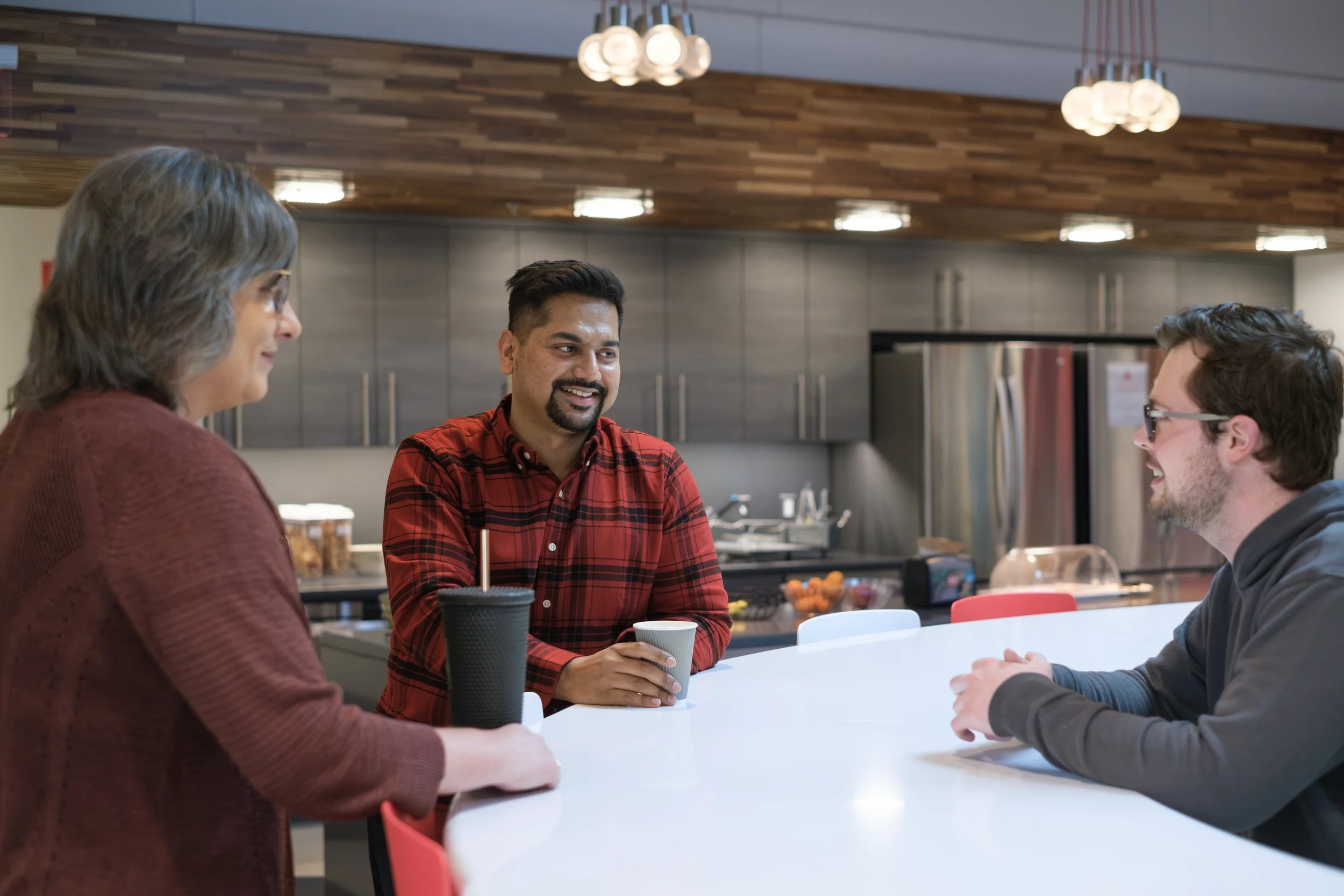 Three people sit at a table while smiling at one another with a coffee cup and water bottle on top of the table.