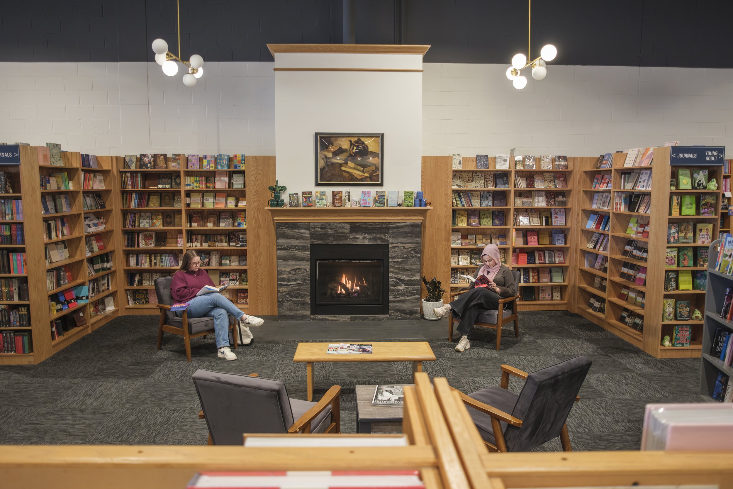 Two people sit in chairs next to a fire place reading books while surrounded by wooden shelves of books.