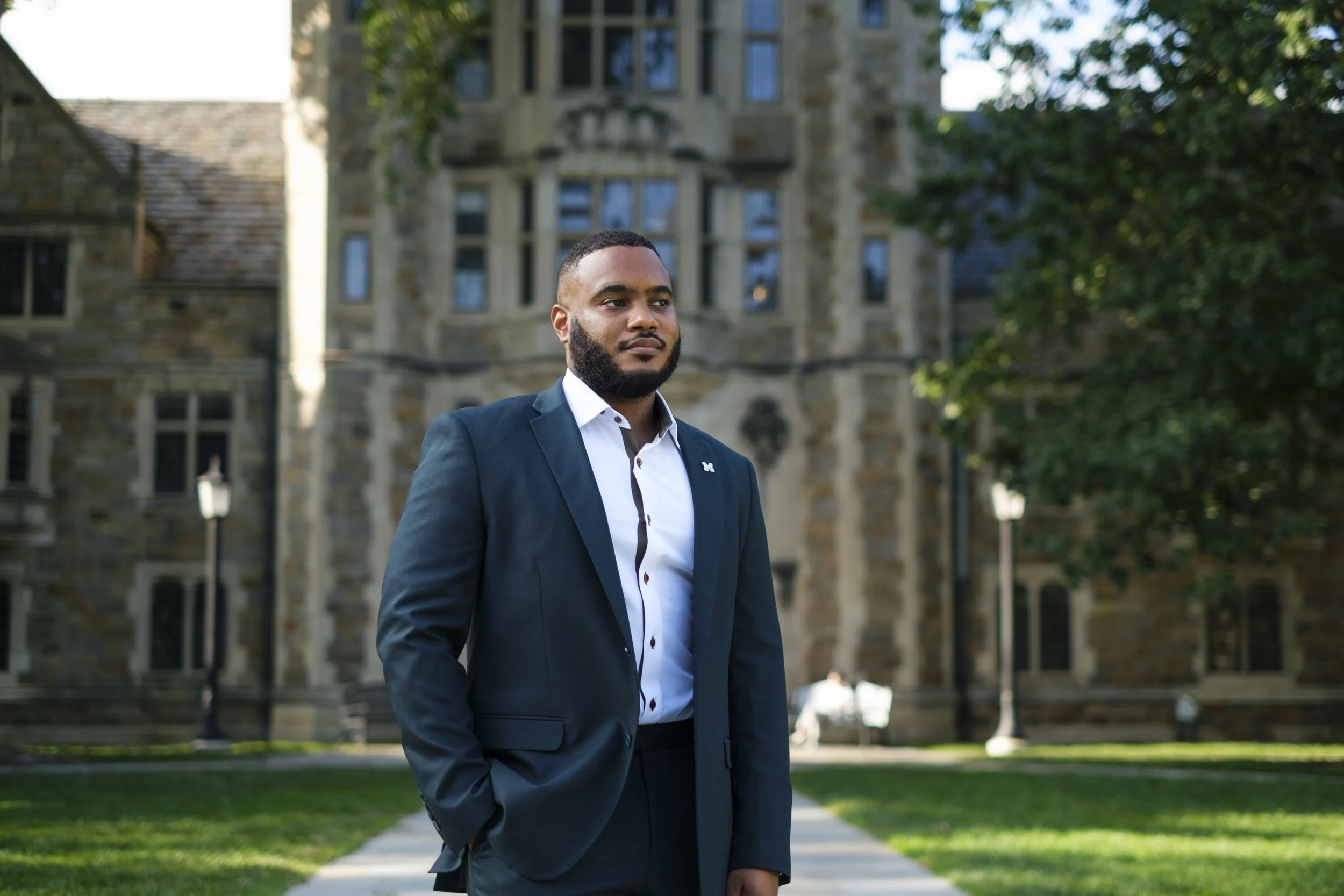 A man stands in a suit while looking off into the distance with the backdrop of the Law Quad at the University of Michigan.