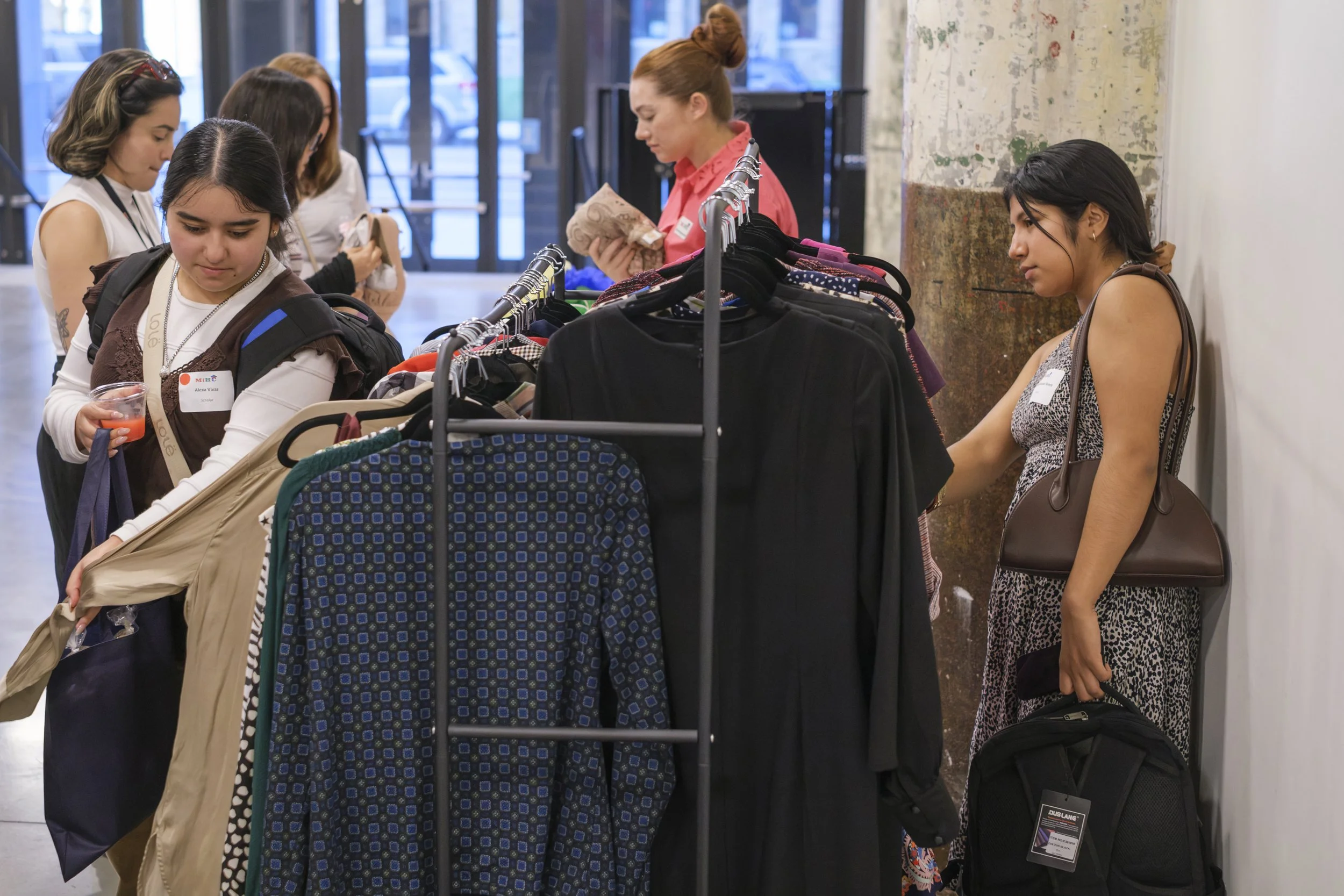 Two young women look through a rack of professional business clothing while holding purses and backpacks during the MiHC Gran Fiesta.