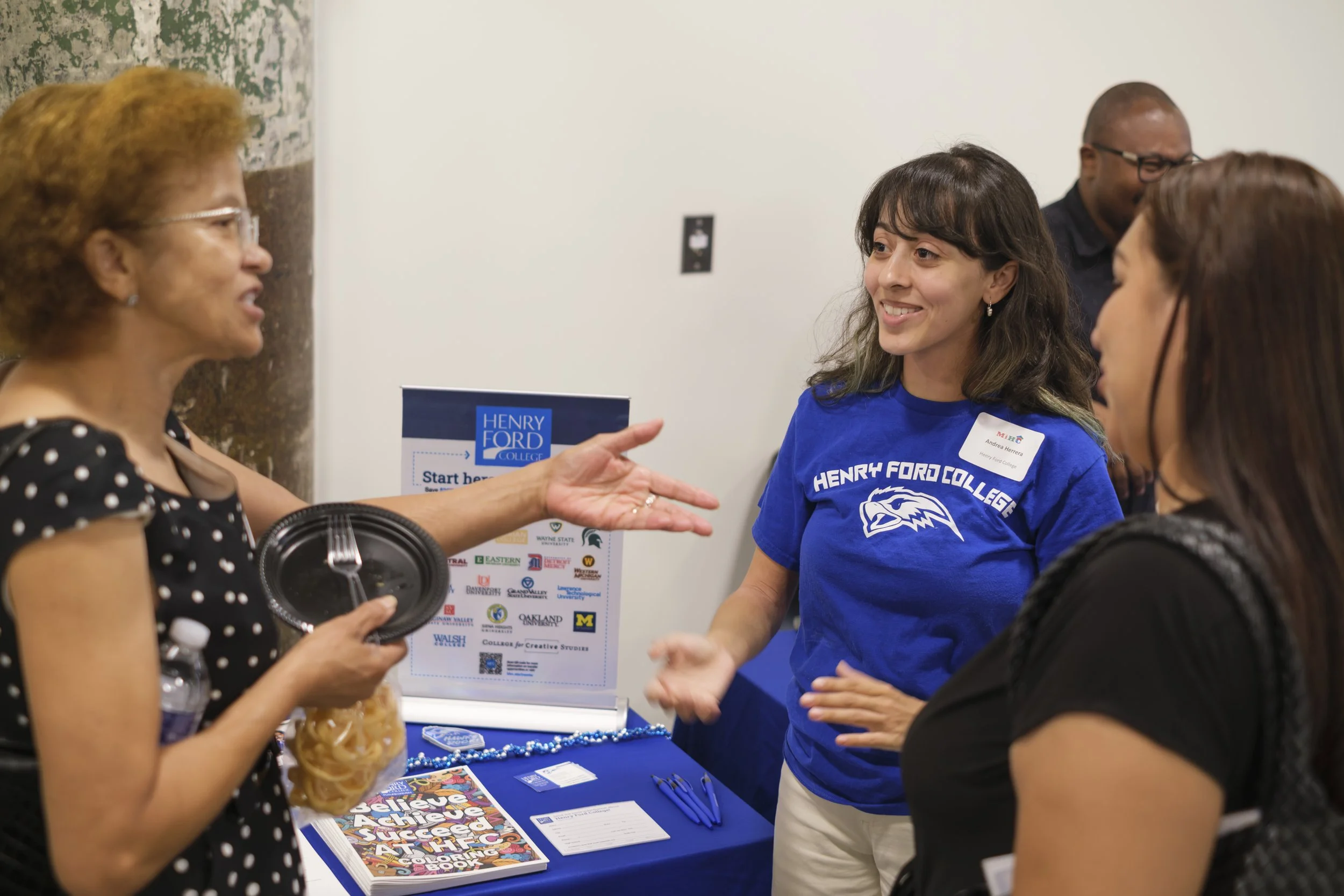 A young woman wearing a Henry Ford College Tshirt smiles while talking to two other people in front of their table.