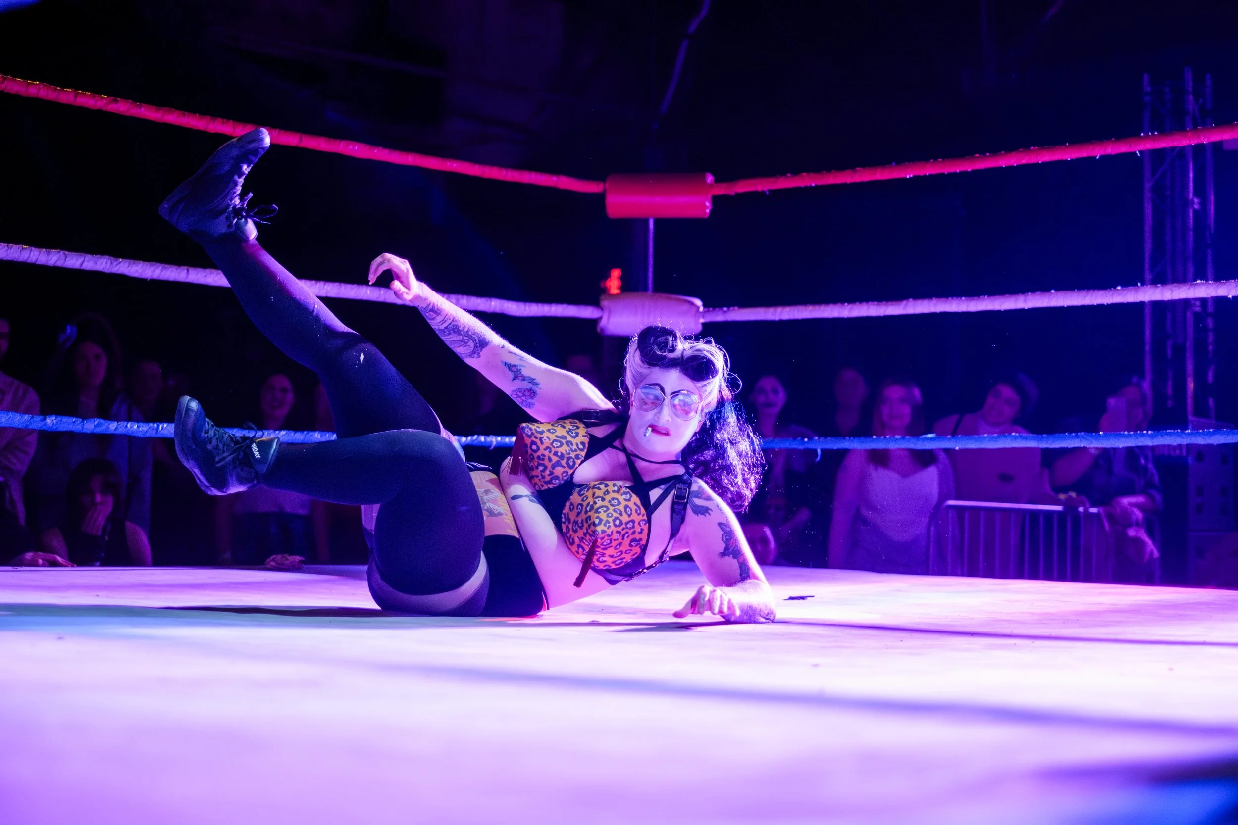 A clown wrestler, known as Rusty Busches, rolls into a wrestling ring on her back while holding a cigarette in her mouth. The stage is lit with purple stage lighting.