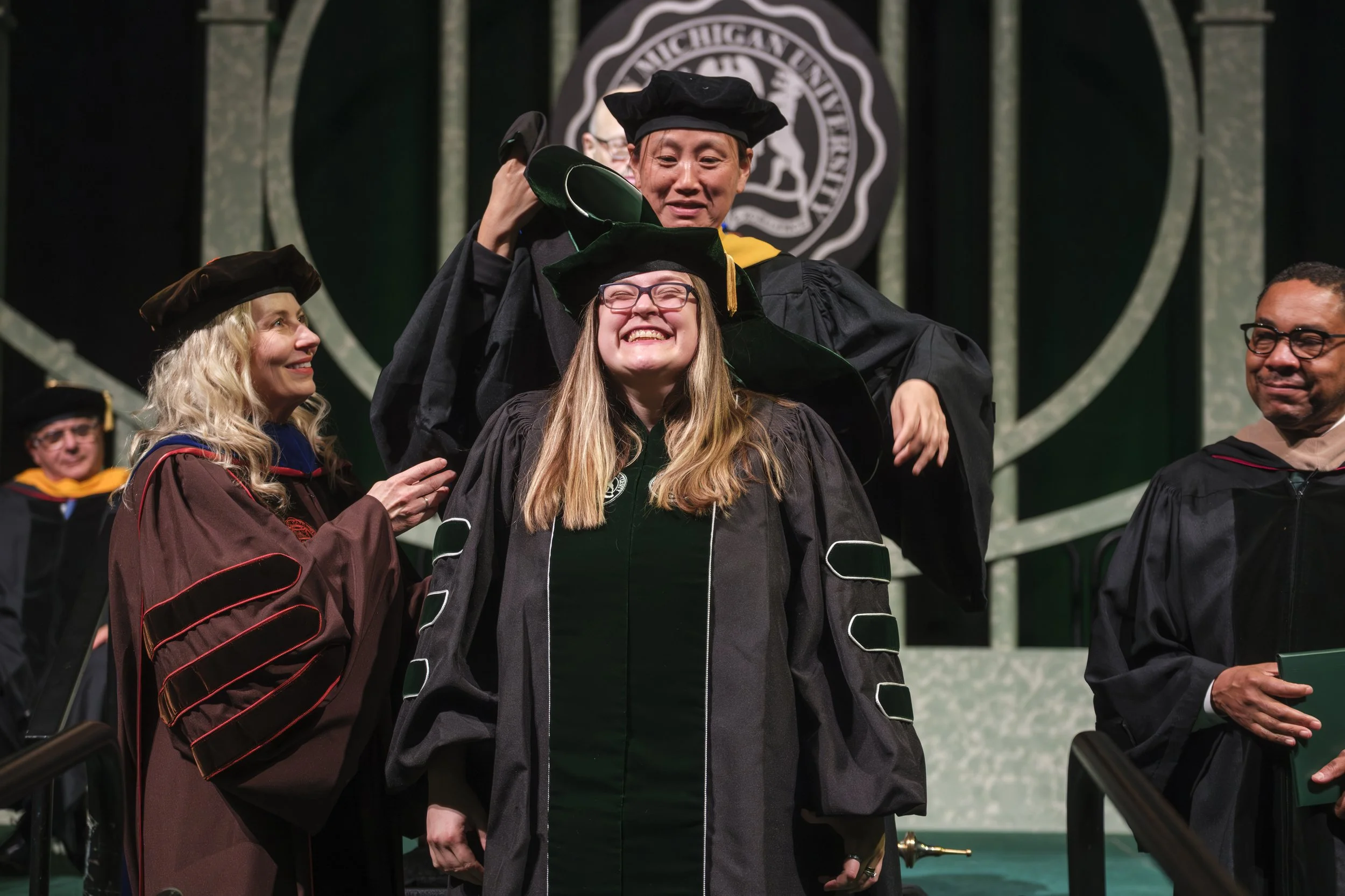 A graduate smiles while receiving their doctoral hood, while someone stands above them, placing it on them and another person stands to the right.