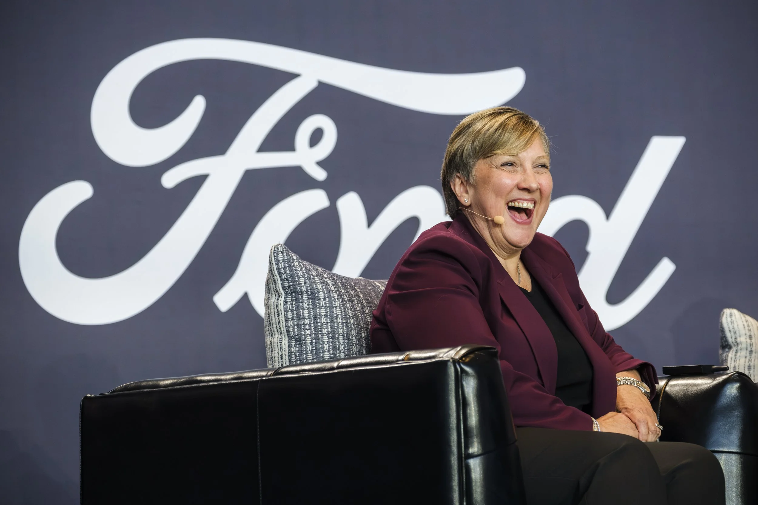 A woman in a magenta suit laughs into a headset microphone while sitting on a black leather chair on stage with a backdrop that reads "Ford" sits behind her.