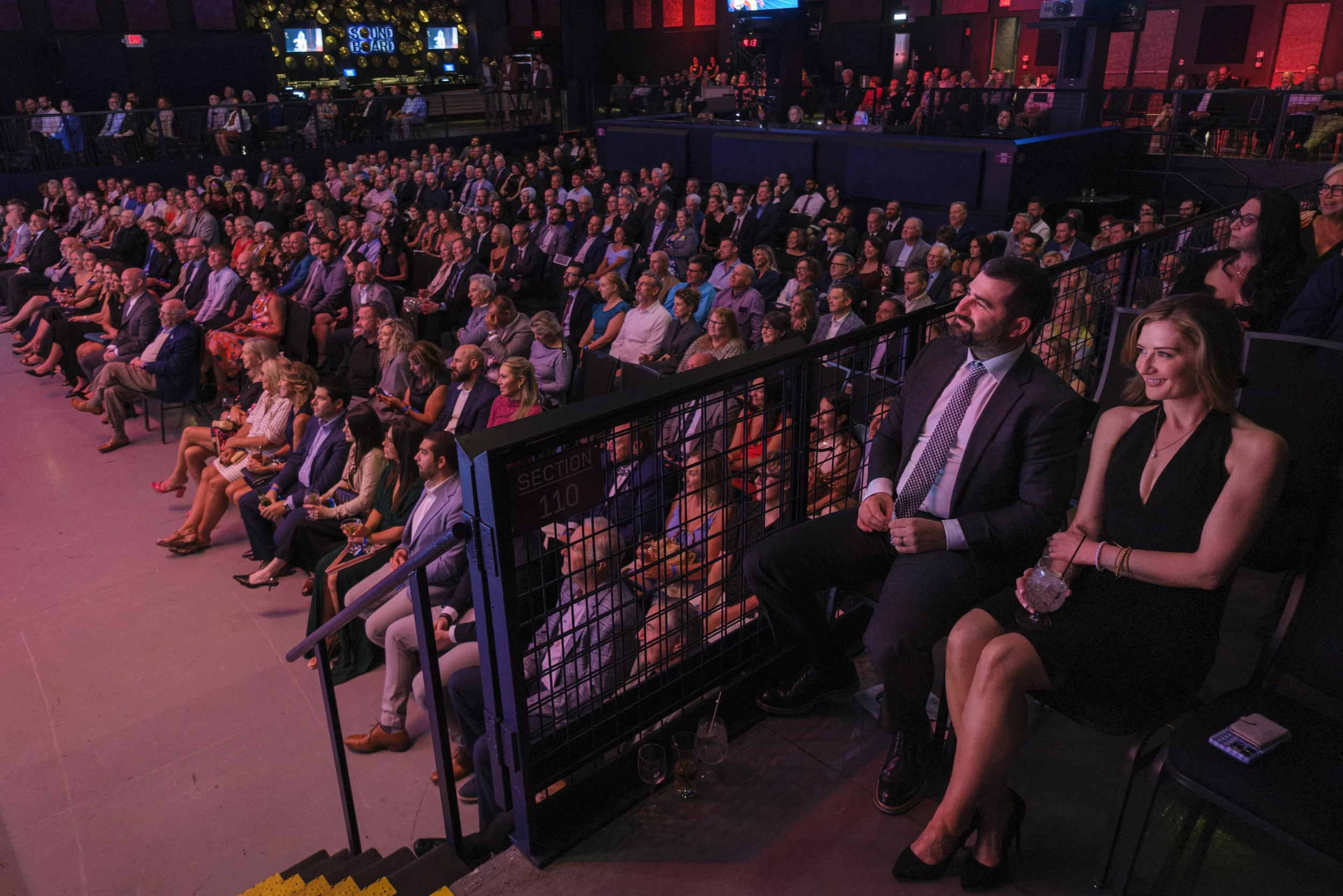 A crowd of people sitting in an audience, dressed in suits and gowns, laugh while watching the roast of Chris Osgood, raising funds for the Jamie Daniels Foundation.