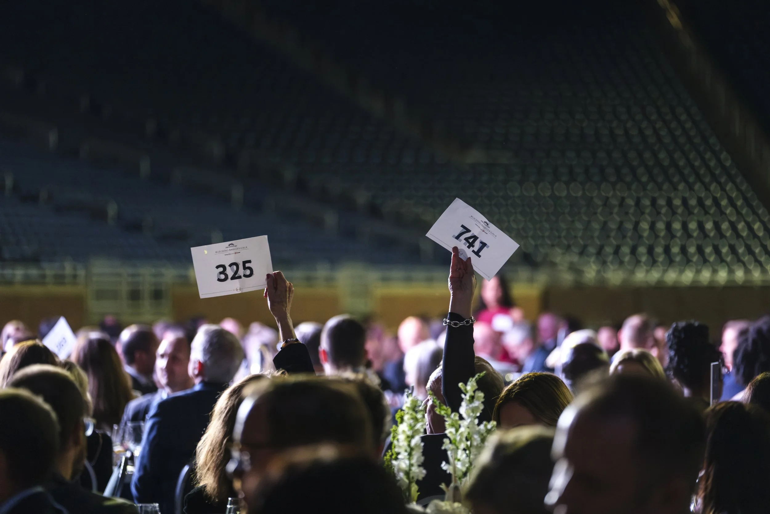 Hands holding up numbers "325" and "741" among a crowd of tables during the fundraising portion of the non profit Pope Francis Center Building Bridges Gala at Ford Field in Detroit.
