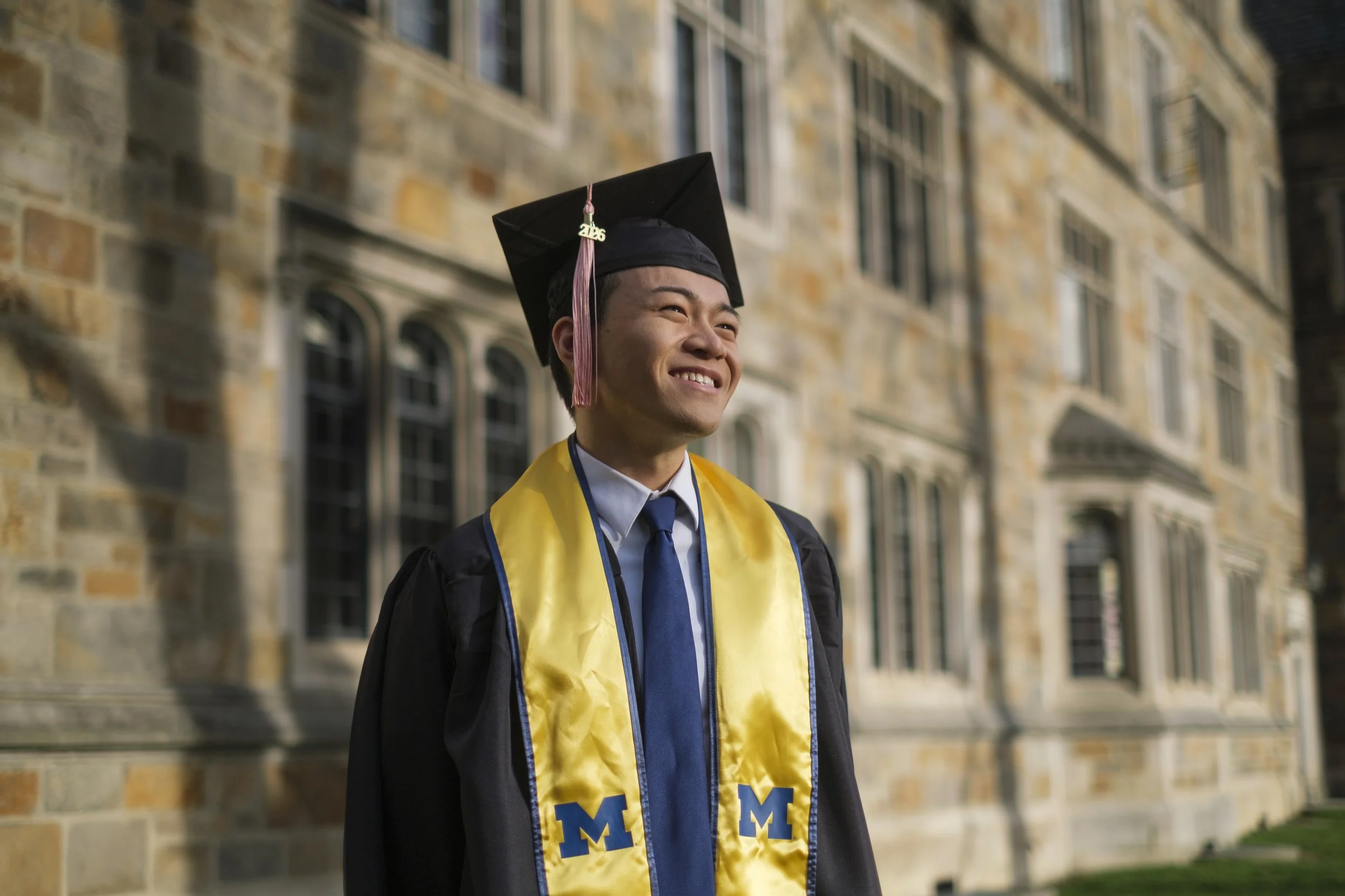 A person smiles while wearing a cap with a tassel and graduation gown with a yellow sash while standing in front of a brick building.