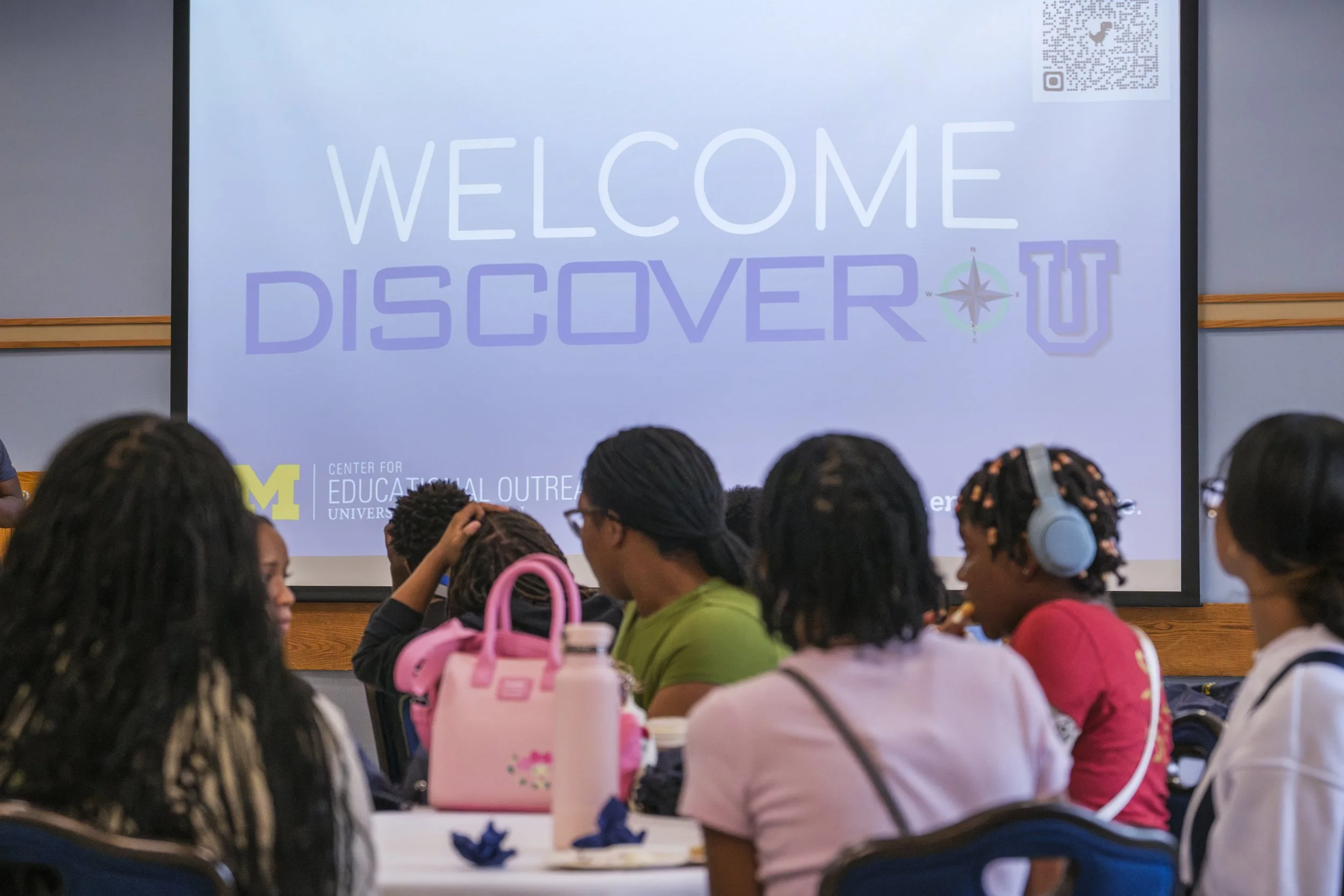 A projector screen that reads "Welcome Discover U" with a block M representing the University of Michigan with students sitting at tables in the foreground.