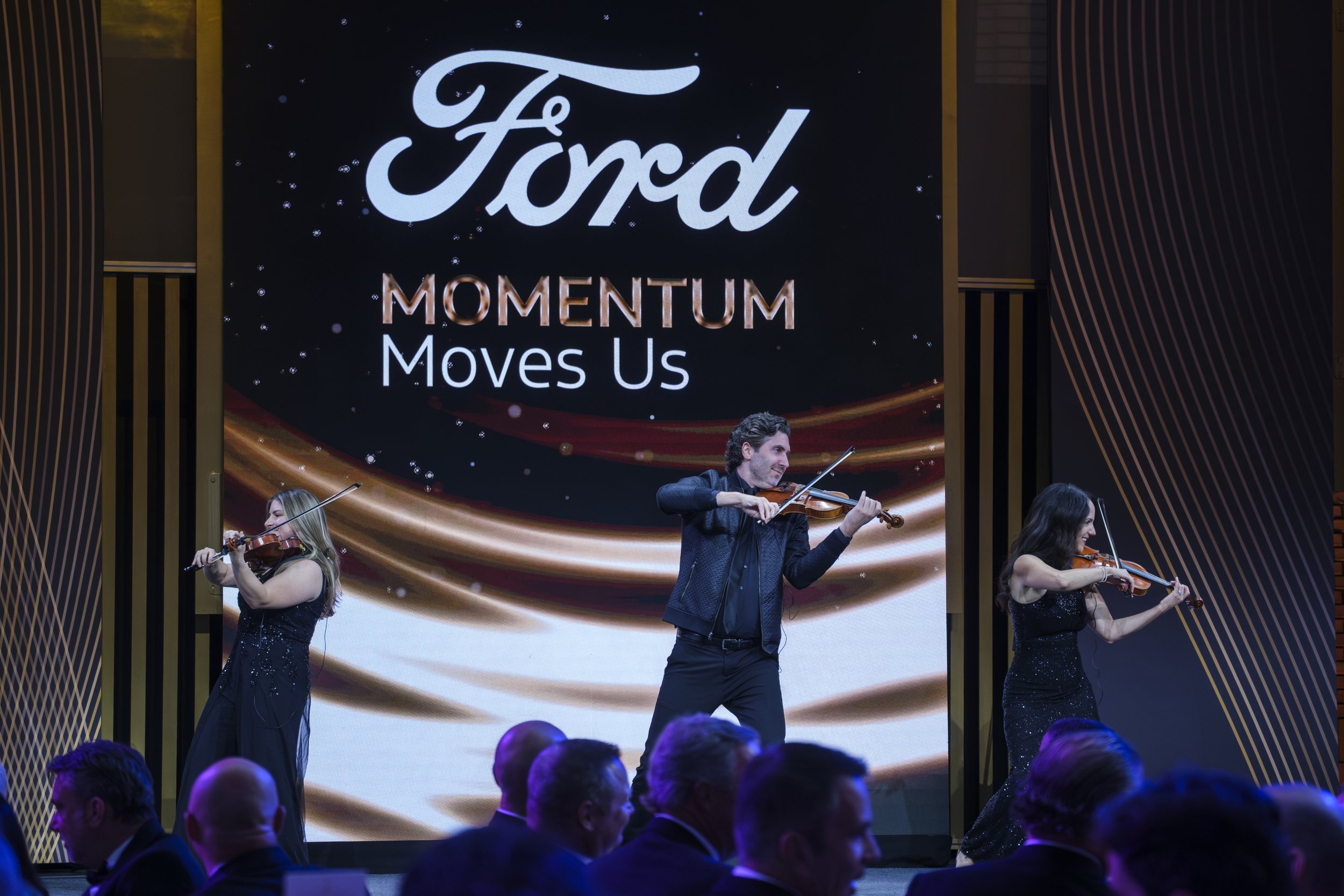A trio of musicians holding stringed instruments perform on stage during the Ford Supplier Summit Awards at Michigan Central Station.